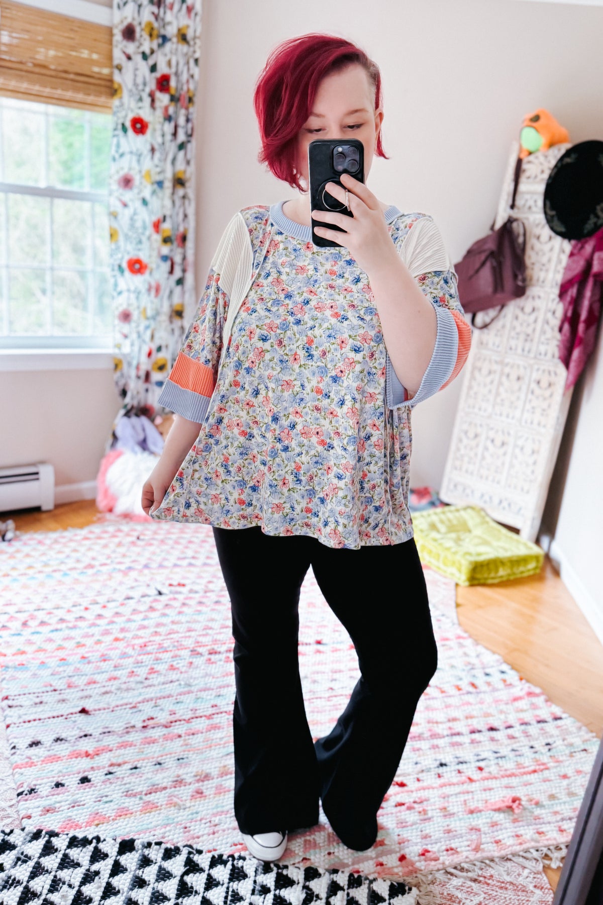 Woman taking a mirror selfie in a floral tunic top with striped sleeves and black pants in a colorful bedroom