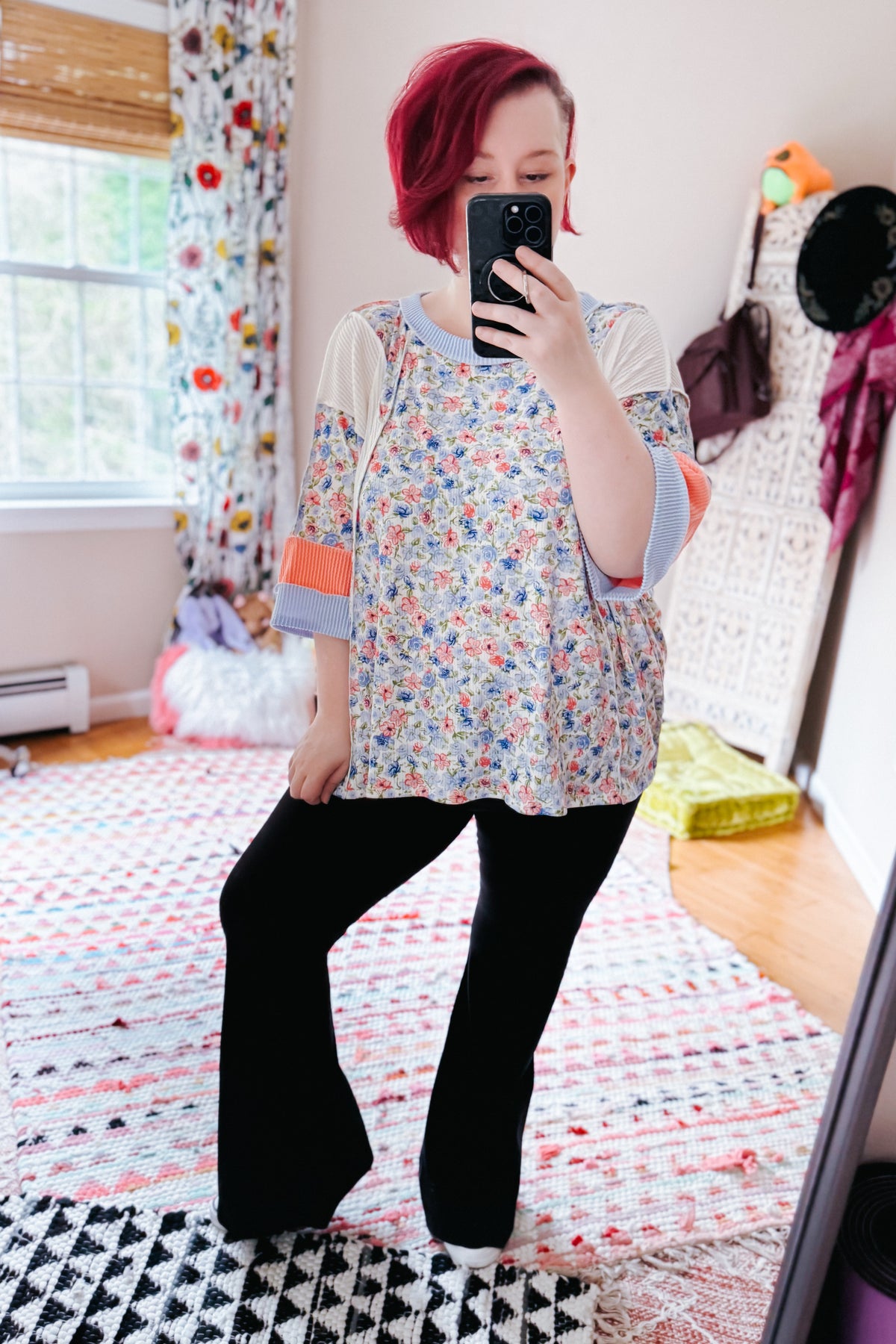 Woman taking a mirror selfie in a blue floral color block top with contrasting sleeves and black flared pants in a bright bedroom