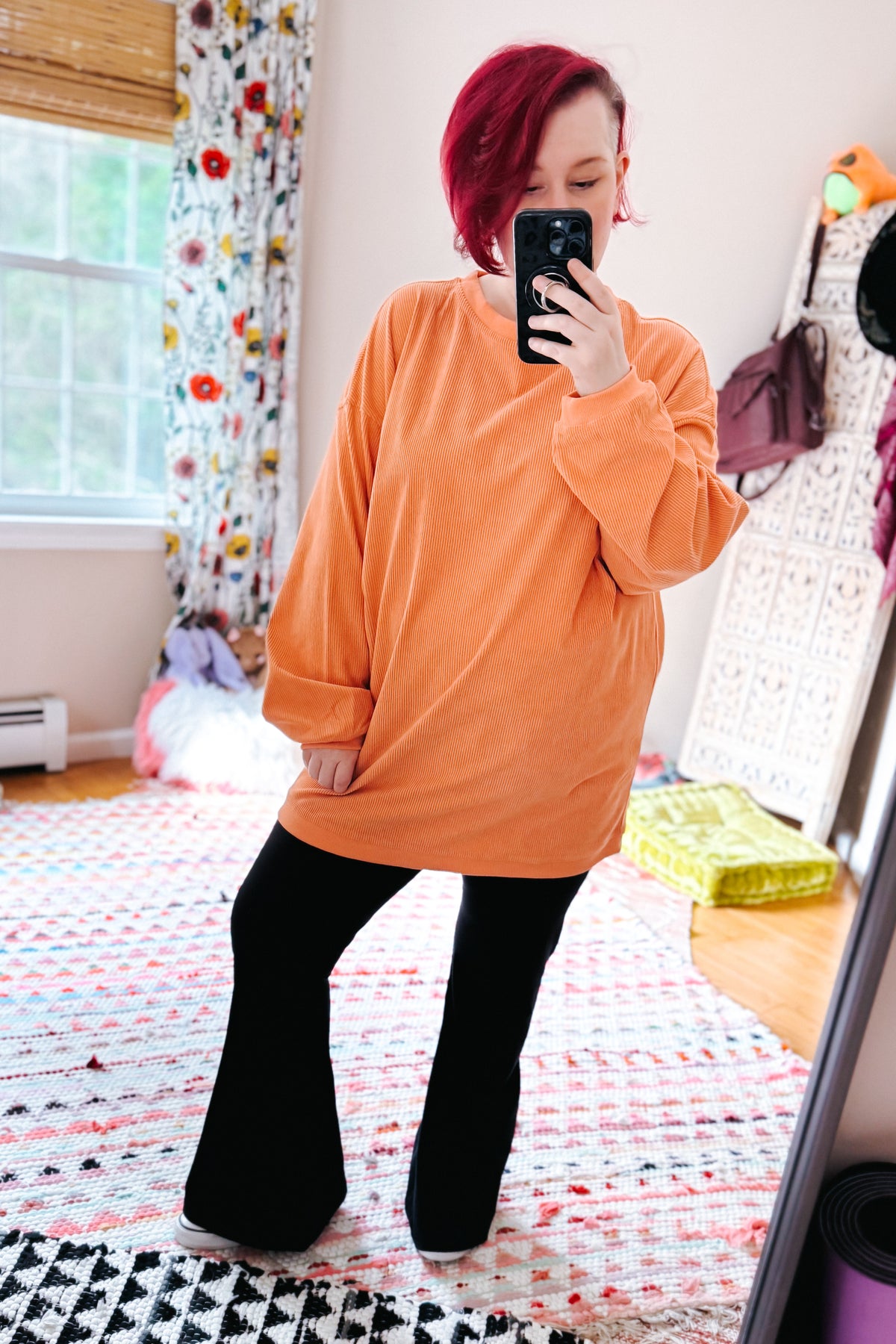 Red haired woman taking a mirror selfie in an oversized orange corduroy sweatshirt and black flared pants in a colorful bedroom
