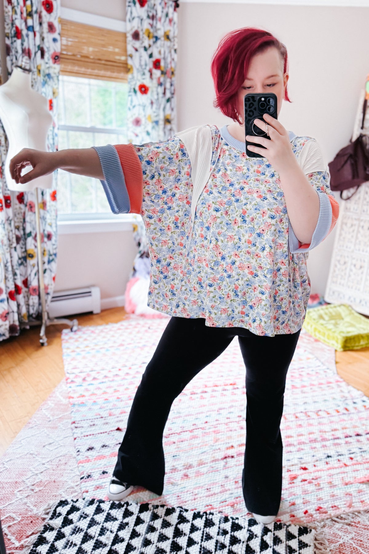 Woman taking a mirror selfie in a blue floral color block knit top with contrasting sleeves and black flared pants in a colorful bedroom