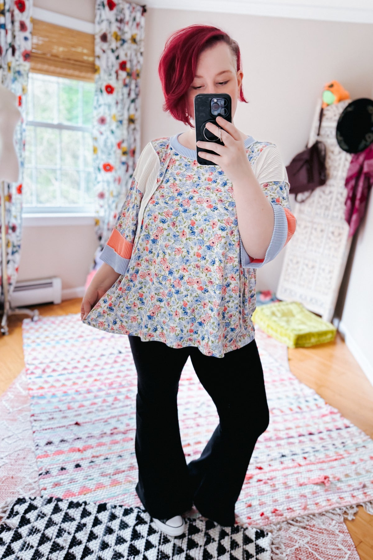 Woman taking a mirror selfie in a blue floral tunic top with patterned sleeves and black pants in a bright bedroom