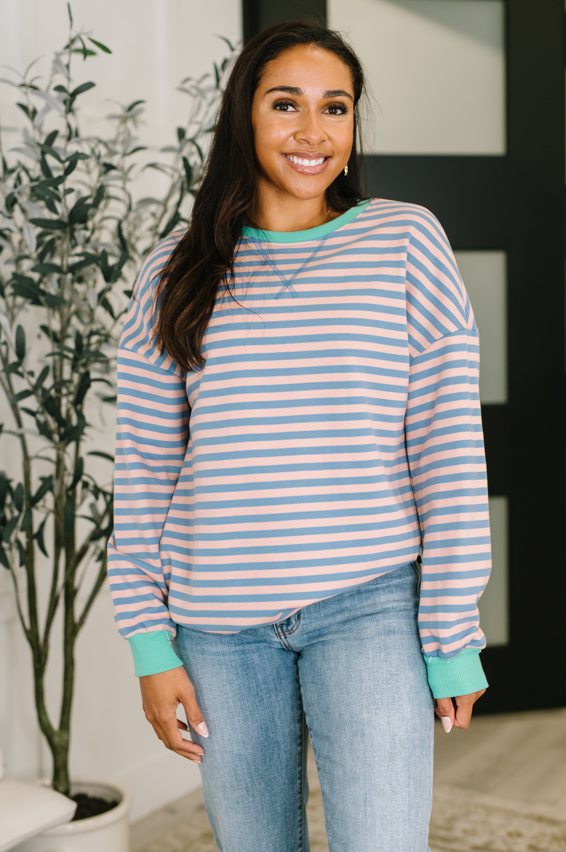 Woman wearing a peach and blue striped drop shoulder pullover and blue jeans standing in an indoor setting.