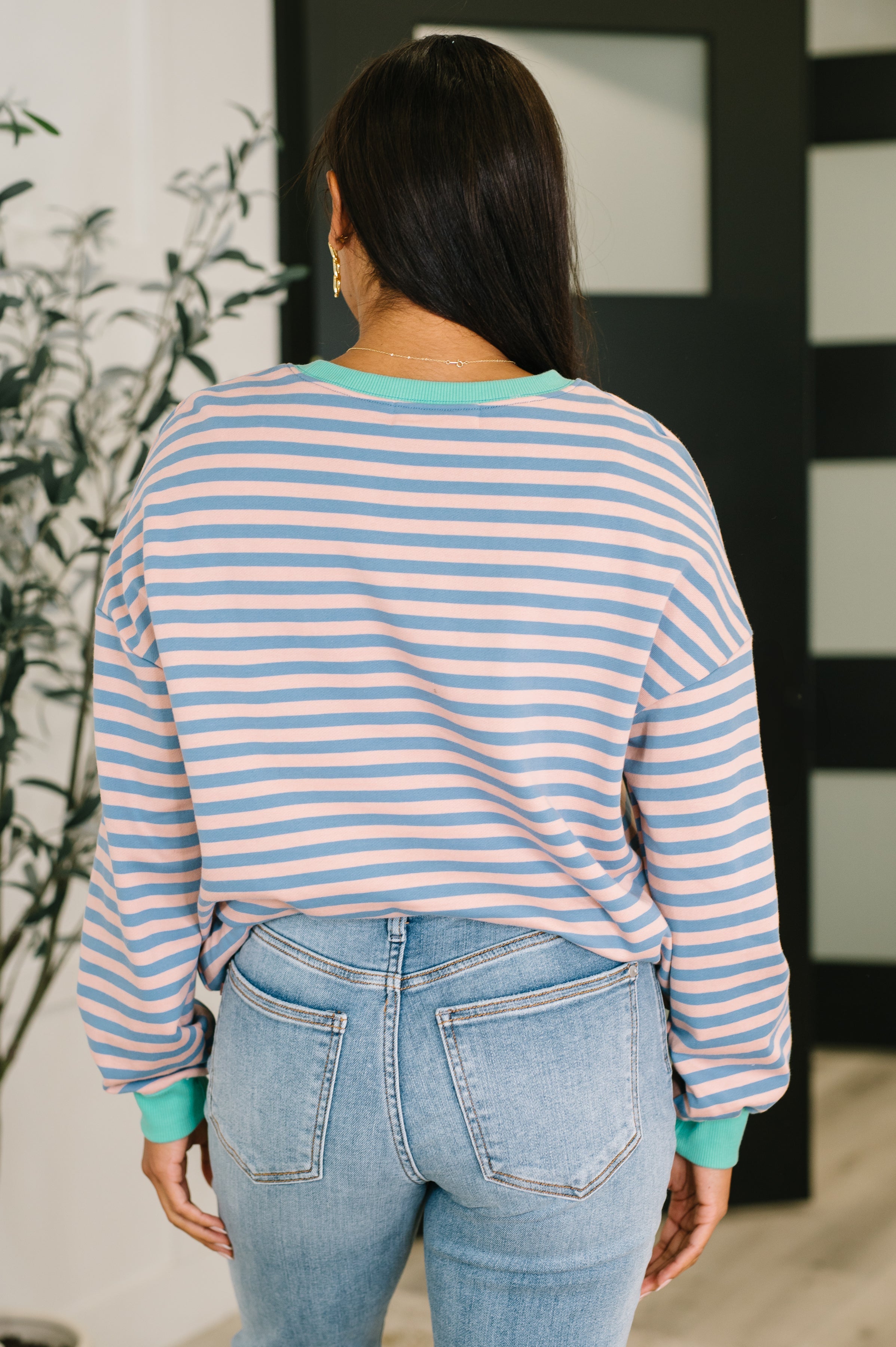 Woman wearing a peach and blue striped drop shoulder pullover and blue jeans standing in an indoor setting.