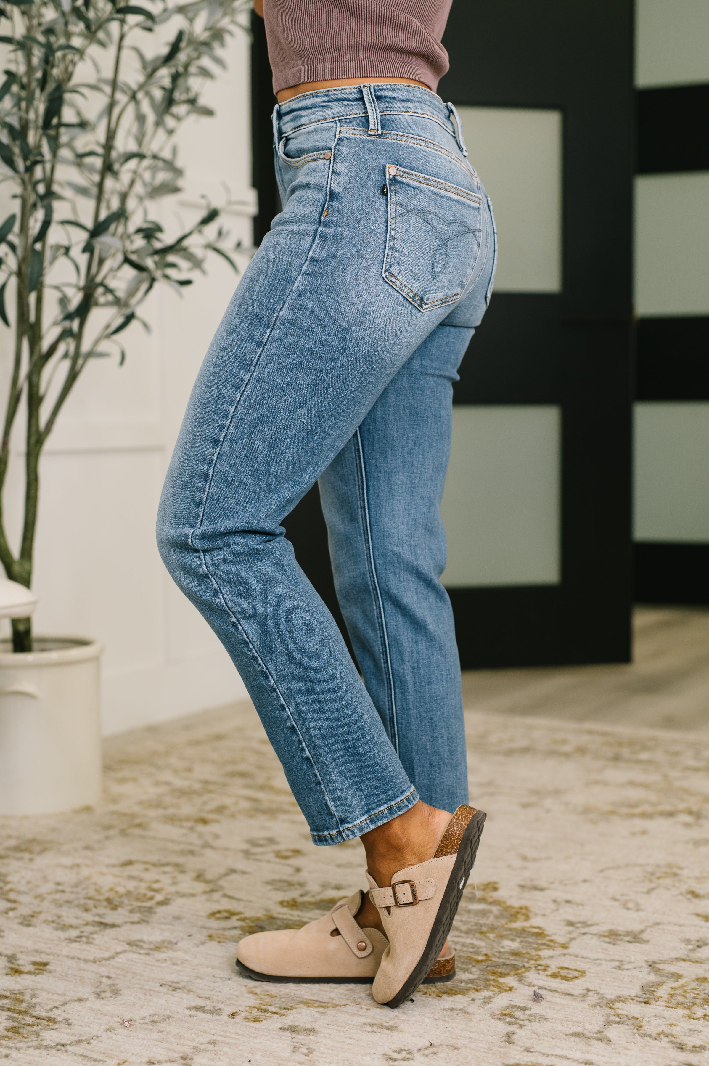 Woman wearing a brown tank top, blue jeans and beige mules standing in an indoor setting.