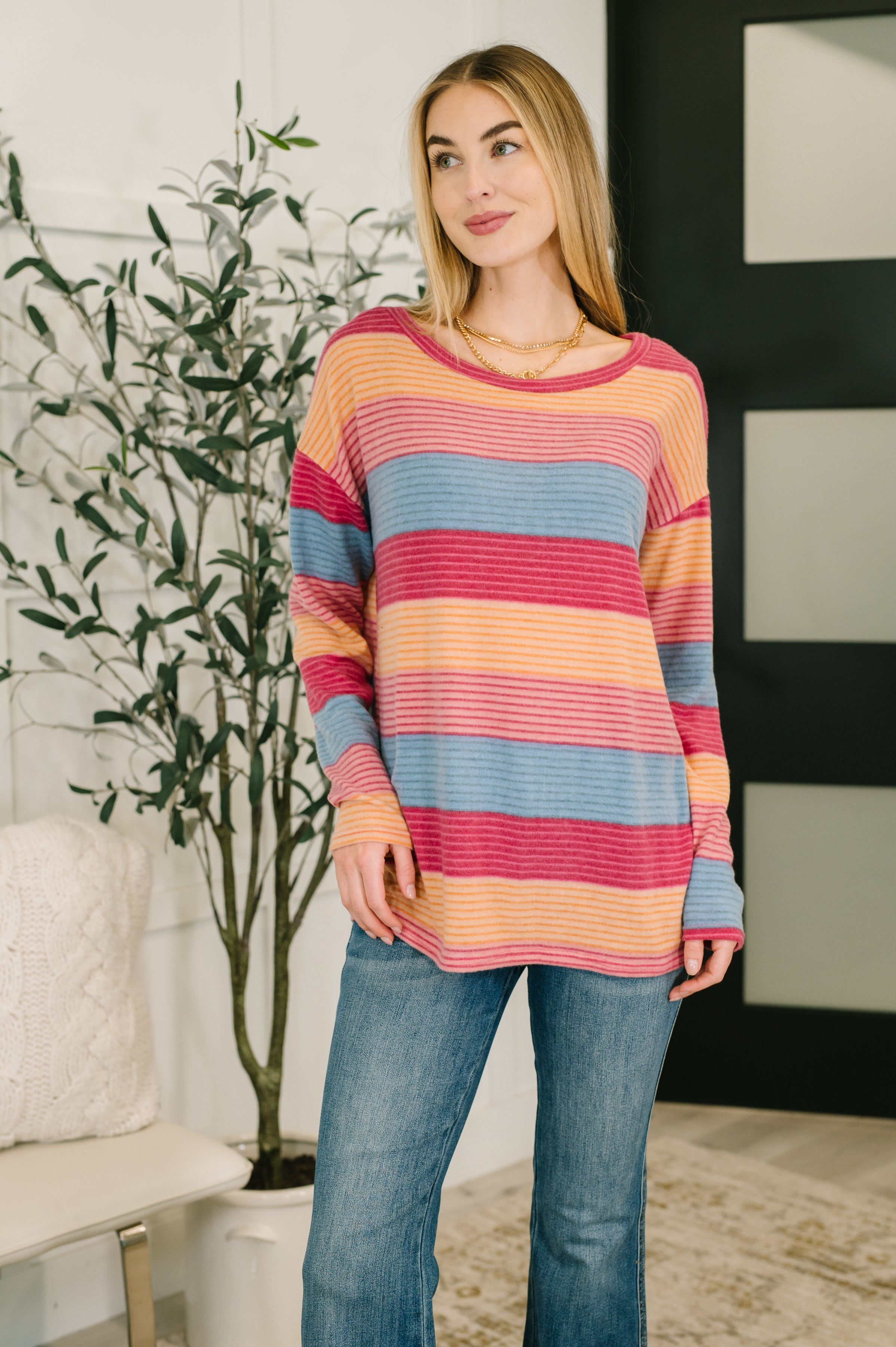 Woman wearing a blue, orange and red striped drop shoulder long sleeve top, a gold necklace and blue jeans in an indoor setting.