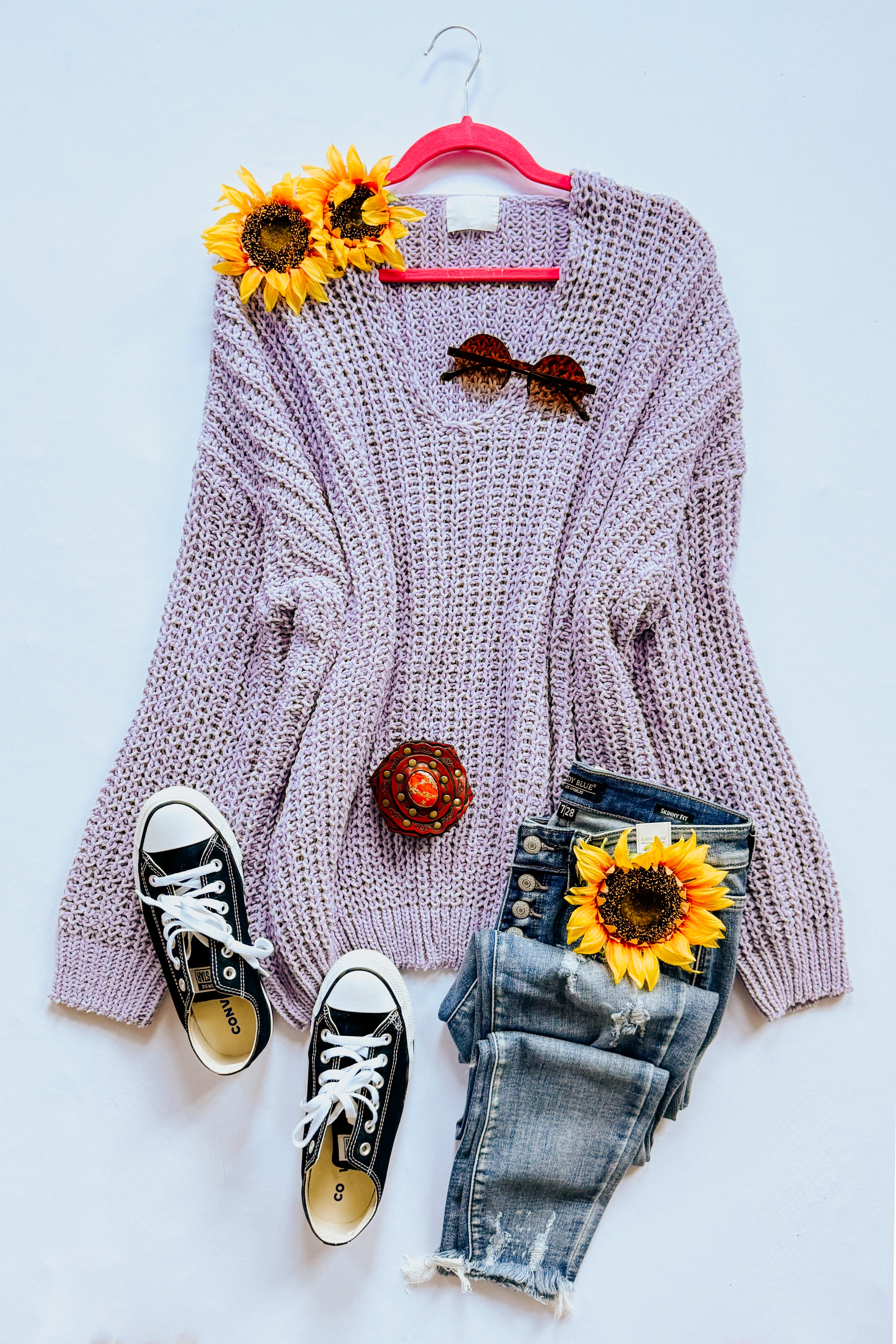 A flat lay setting on a white background of a chunky lavender sweater, blue jeans, converse sneakers, accessories and flowers.