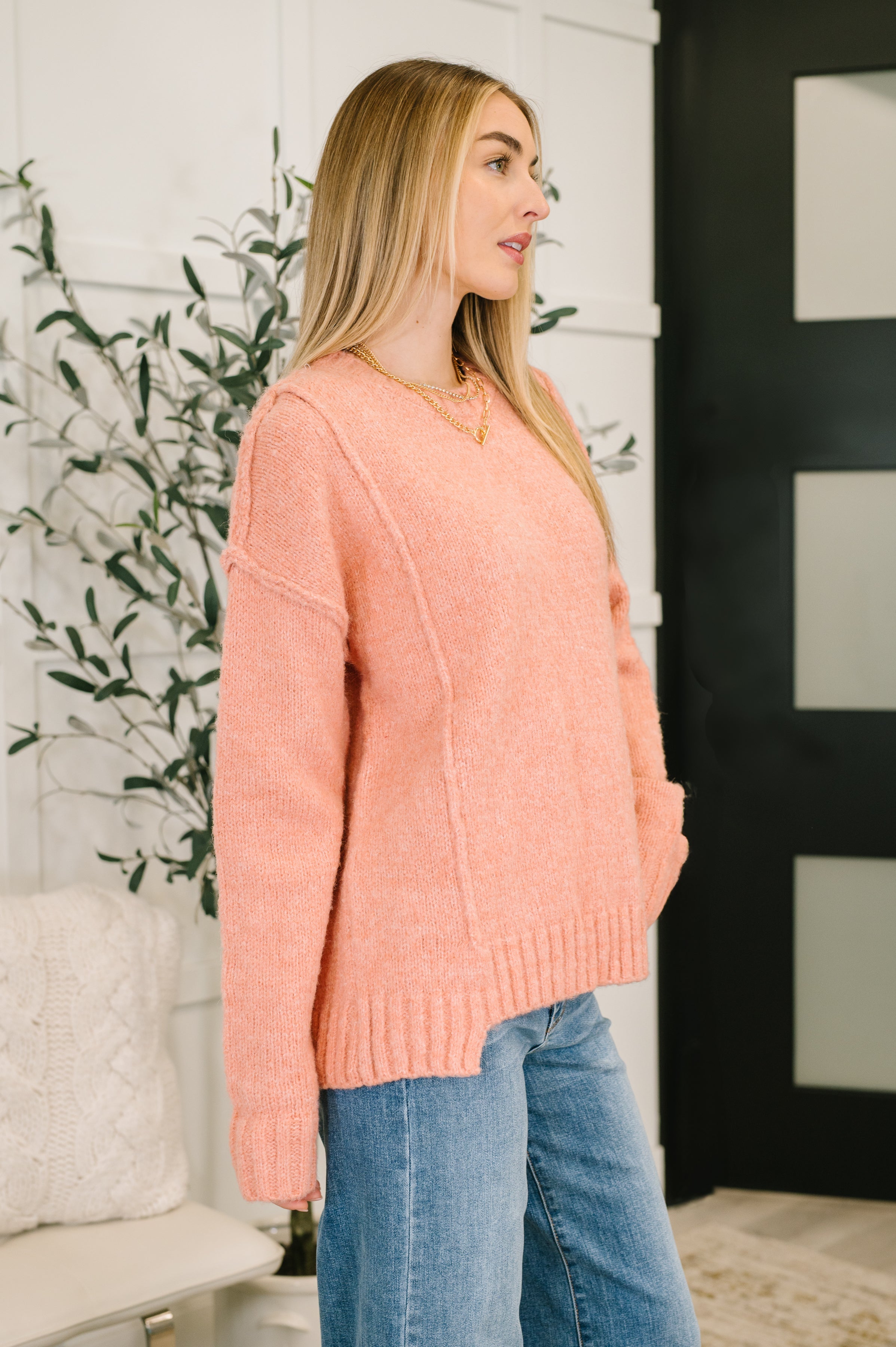 Woman wearing a peach exposed seam long sleeve sweater, a gold necklace and blue jeans in an indoor setting.