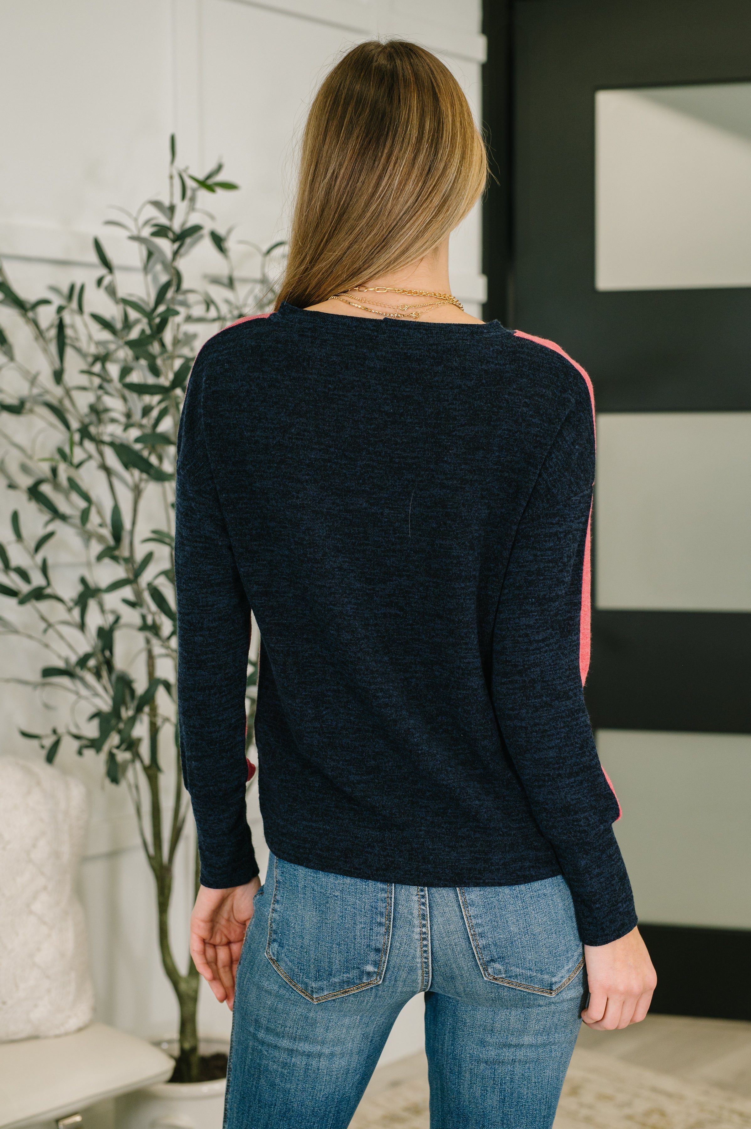 Woman with her back facing the camera wearing a red sweater with black accents and blue jeans, standing in a room with decorative elements.
