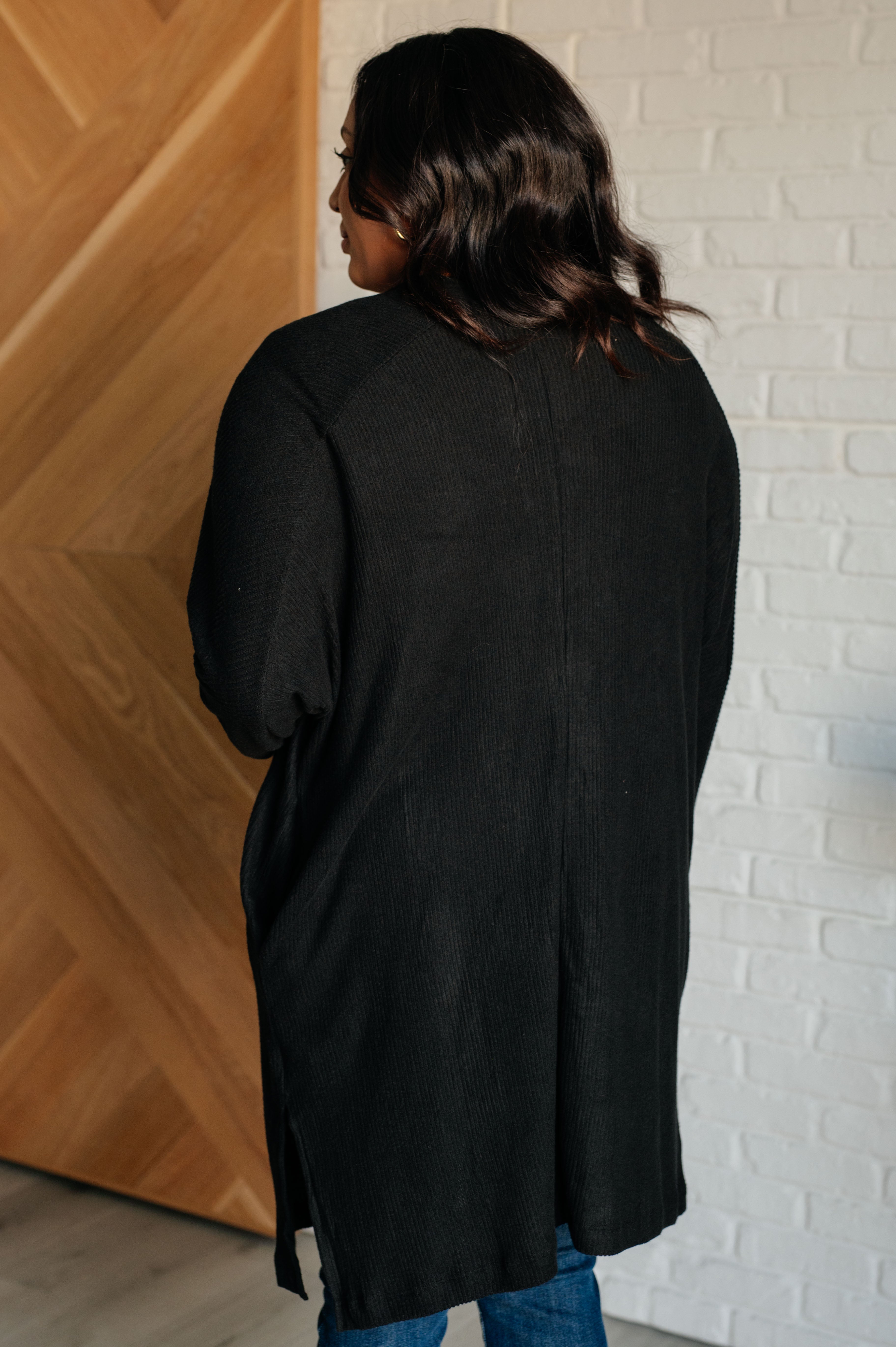 A women wearing a black cardigan standing against a white brick wall.