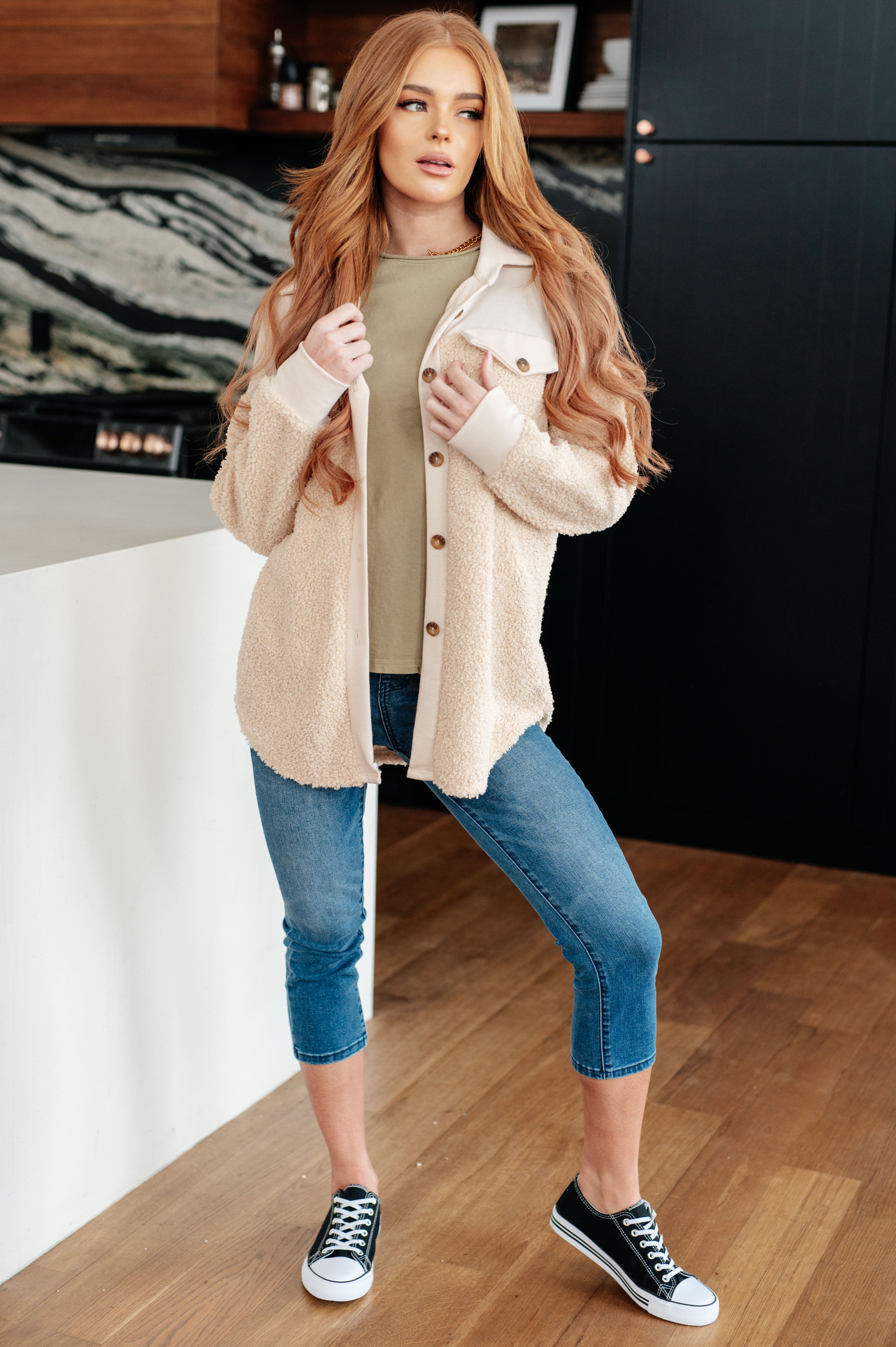 Woman wearing a beige shacket, a green top, blue jeans and tennis shoes in an indoor setting