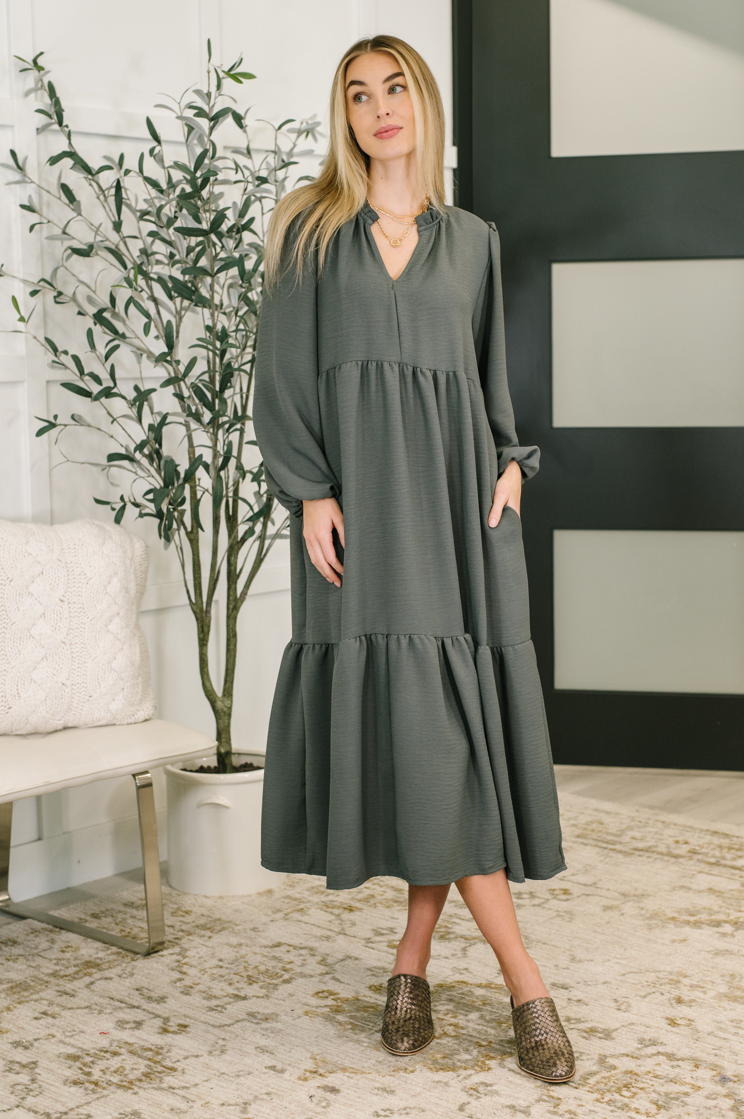 Woman wearing a grey tiered relaxed fit dress standing in a room with a plant and a black and white wall.