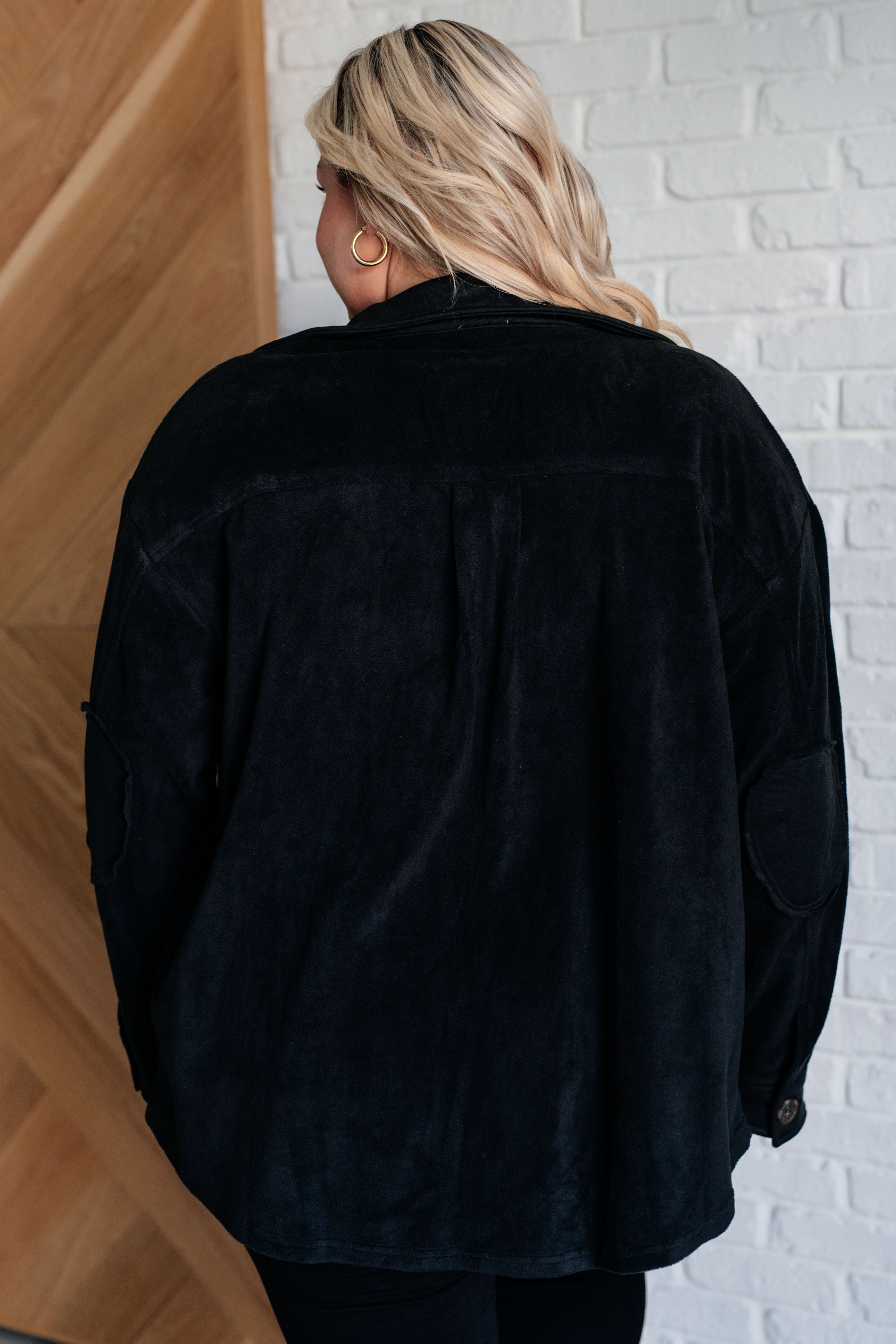 Woman wearing a black fleece button up jacket and black pants standing against a white brick wall.