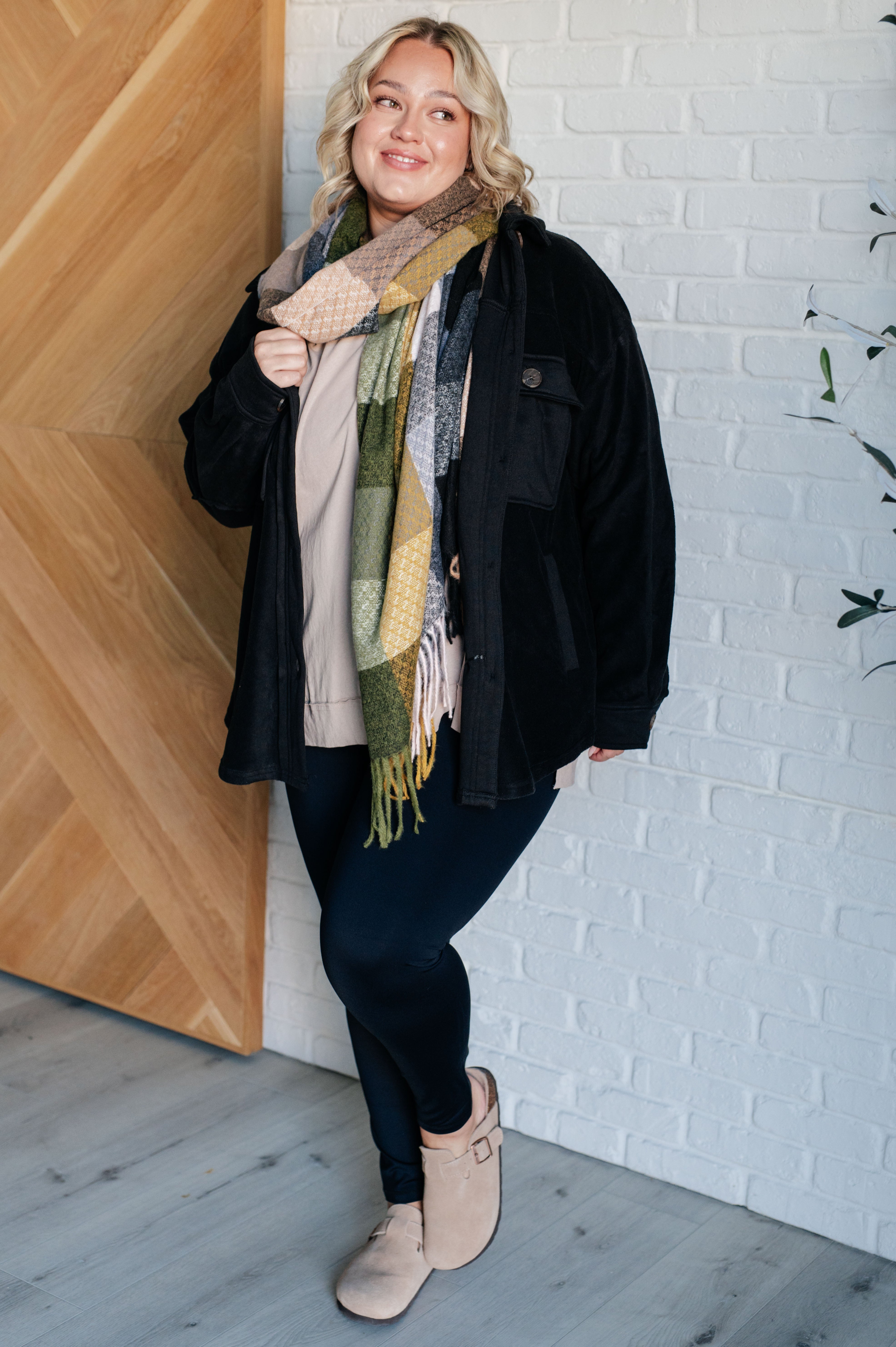 Woman wearing a black fleece button up jacket, a beige top, a green scarf, black pants and tan clogs standing against a white brick wall.