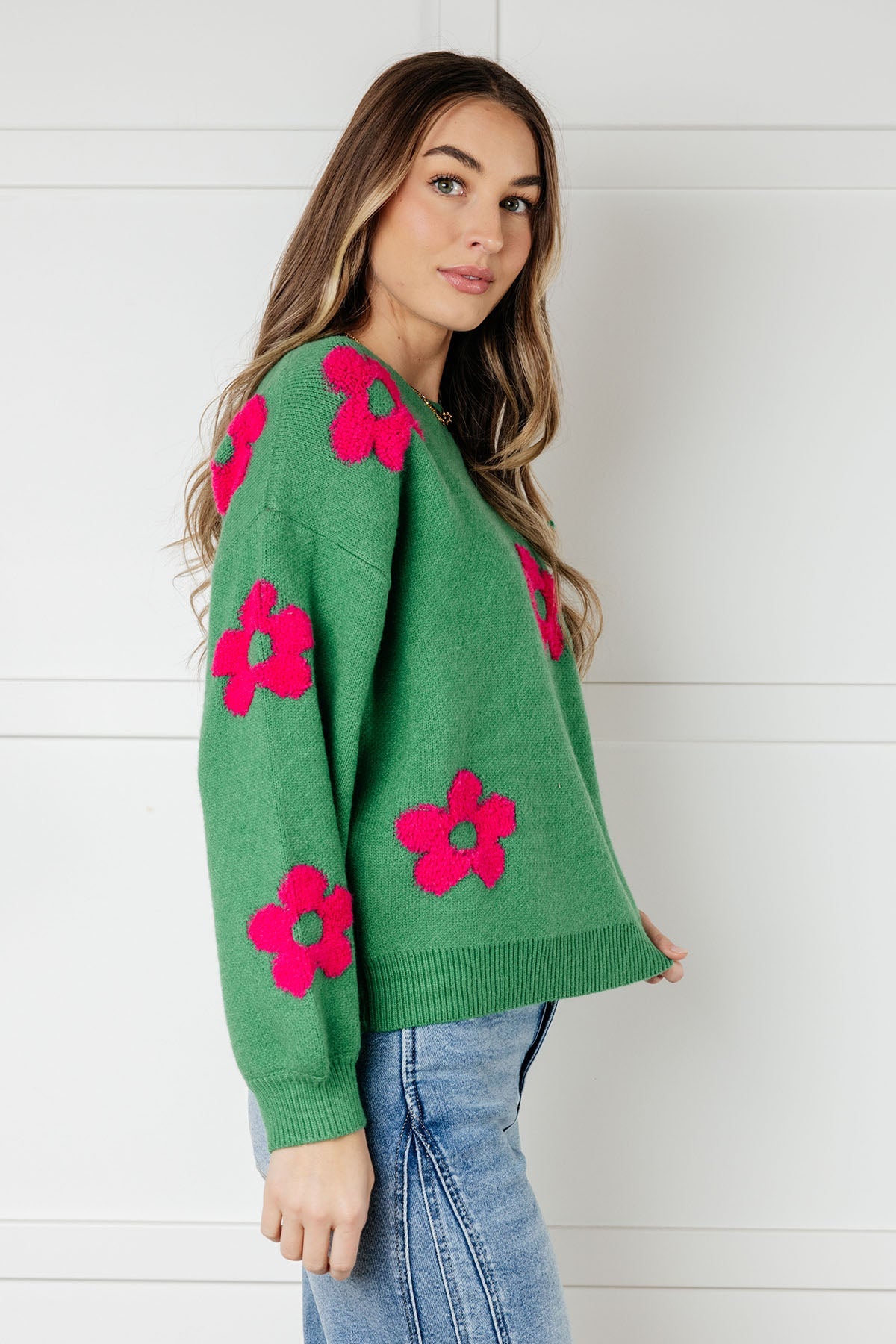 Woman wearing a green sweater embellished with tufted pink flowers and blue jeans against a white wall.