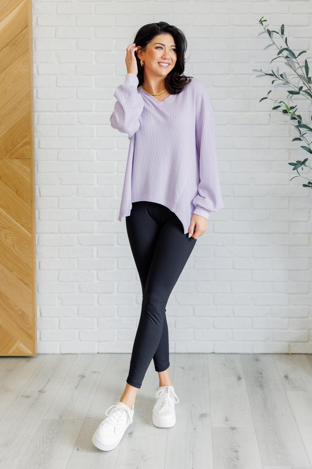Woman wearing a lavender waffle knit v neck long sleeve top, black leggings and white sneakers standing against a white brick wall.