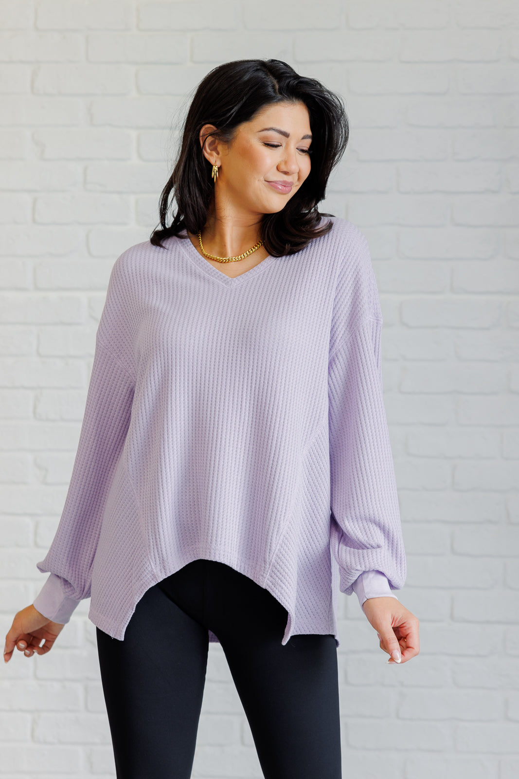 Woman wearing a lavender waffle knit v neck long sleeve top and black leggings standing against a white brick wall.