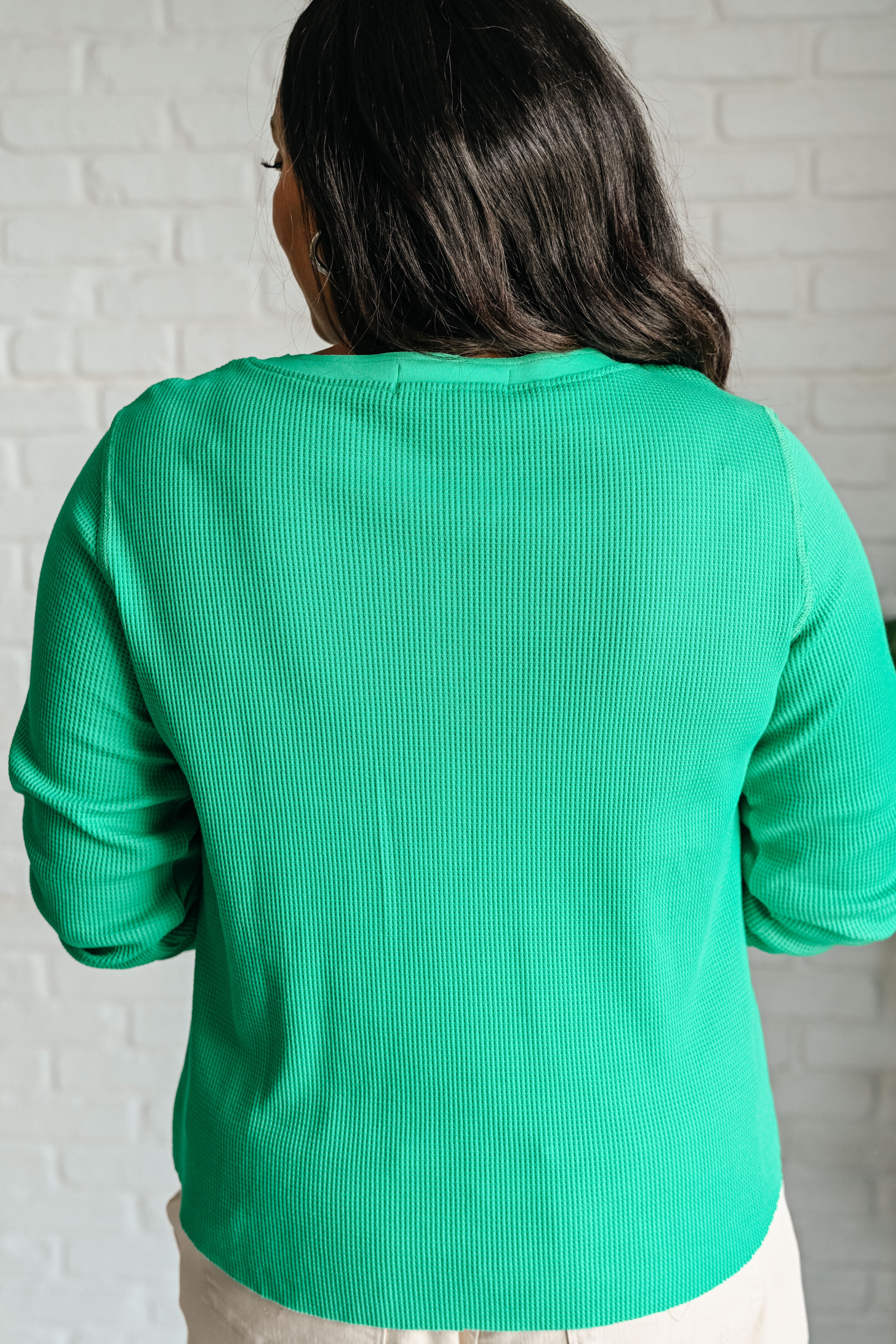 Woman wearing a green long-sleeve waffle knit henley top and beige pants with a white brick wall background.
