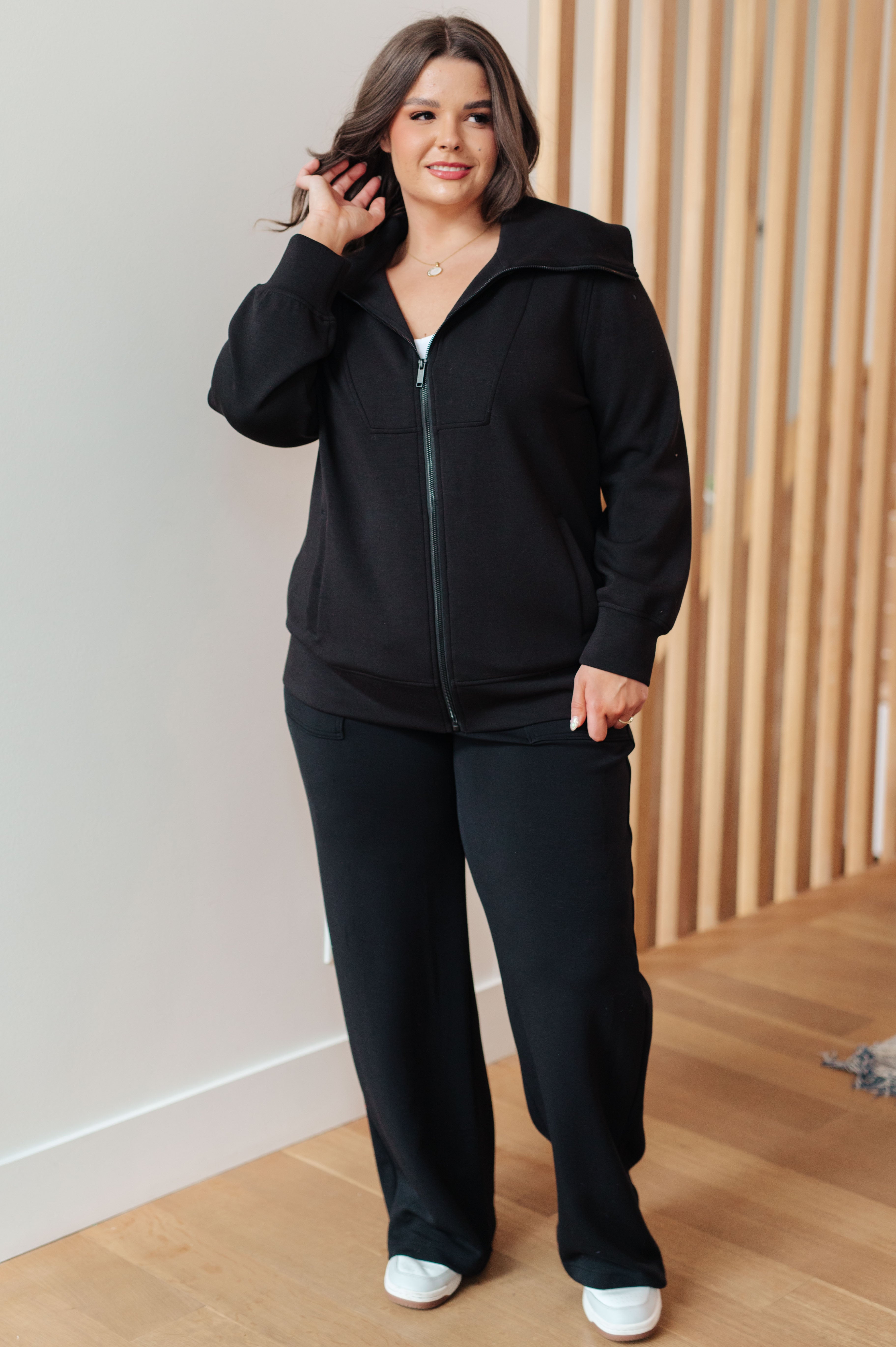 Woman wearing a athleisure black high neck zip up jacket, black pants and white sneakers standing in an indoor setting.