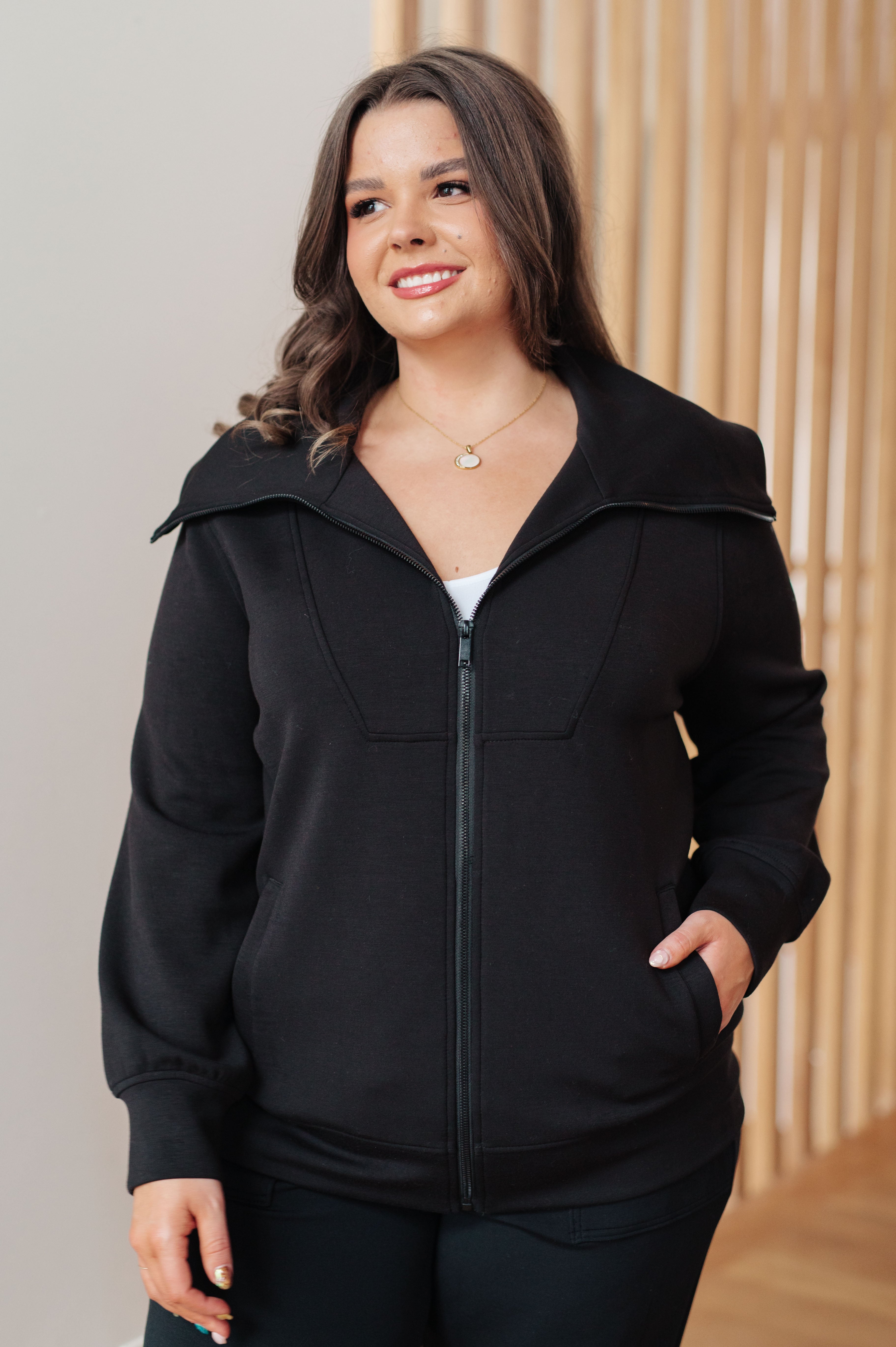 Woman wearing a athleisure black high neck zip up jacket and black pants standing in an indoor setting.