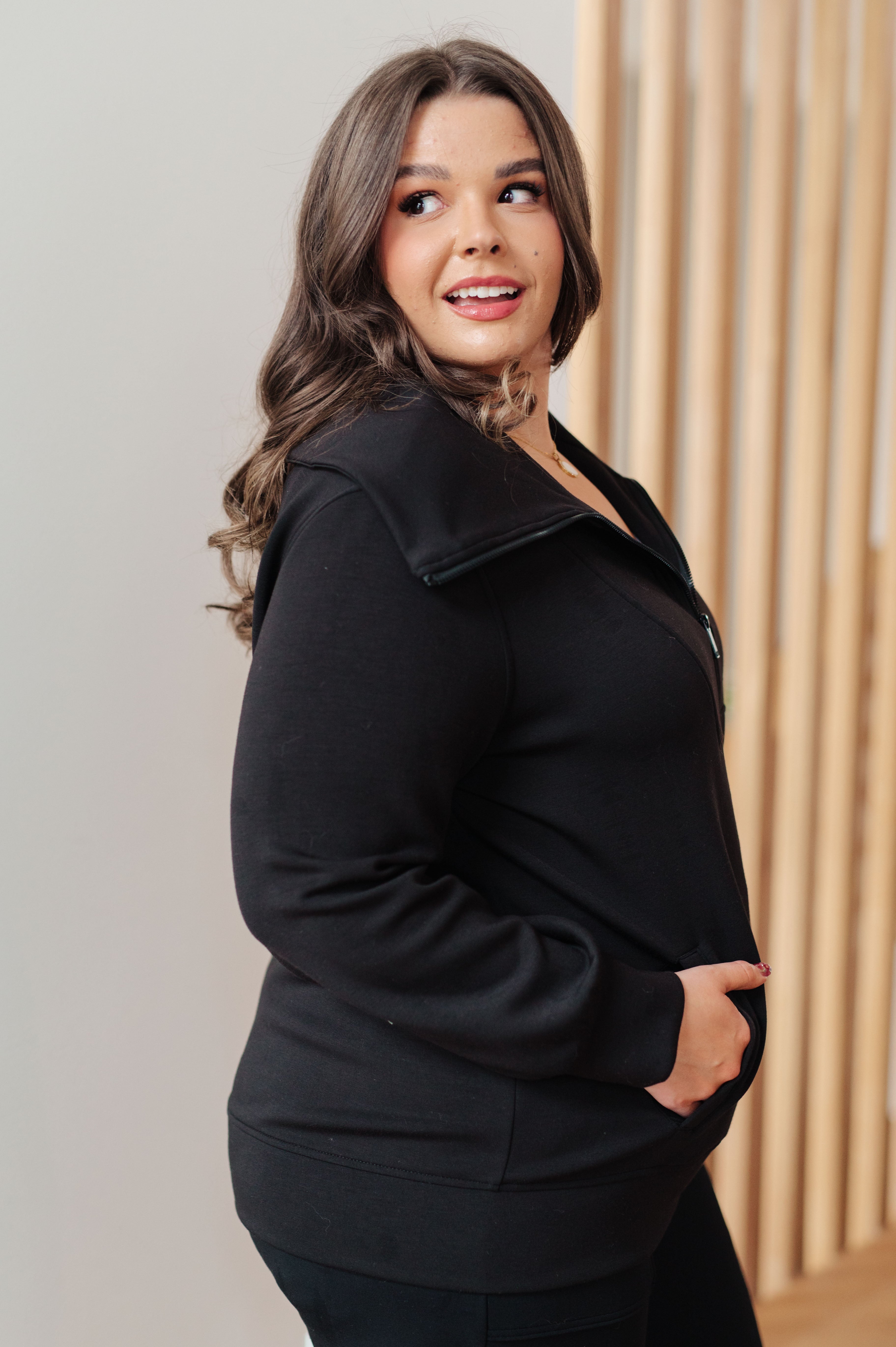 Woman wearing a athleisure black high neck zip up jacket and black pants standing in an indoor setting.