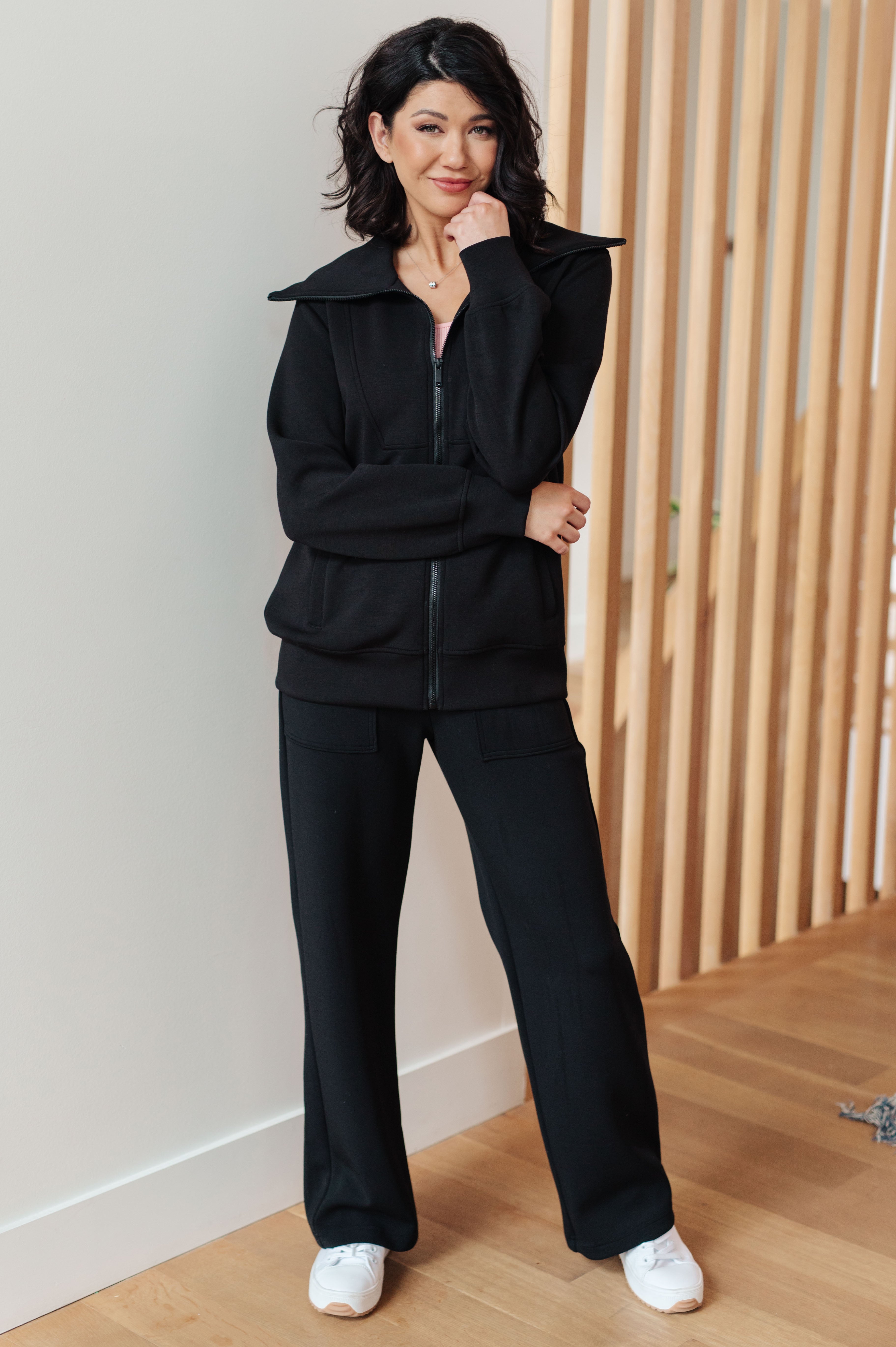 Woman wearing a athleisure black high neck zip up jacket, black pants and white sneakers standing in an indoor setting.