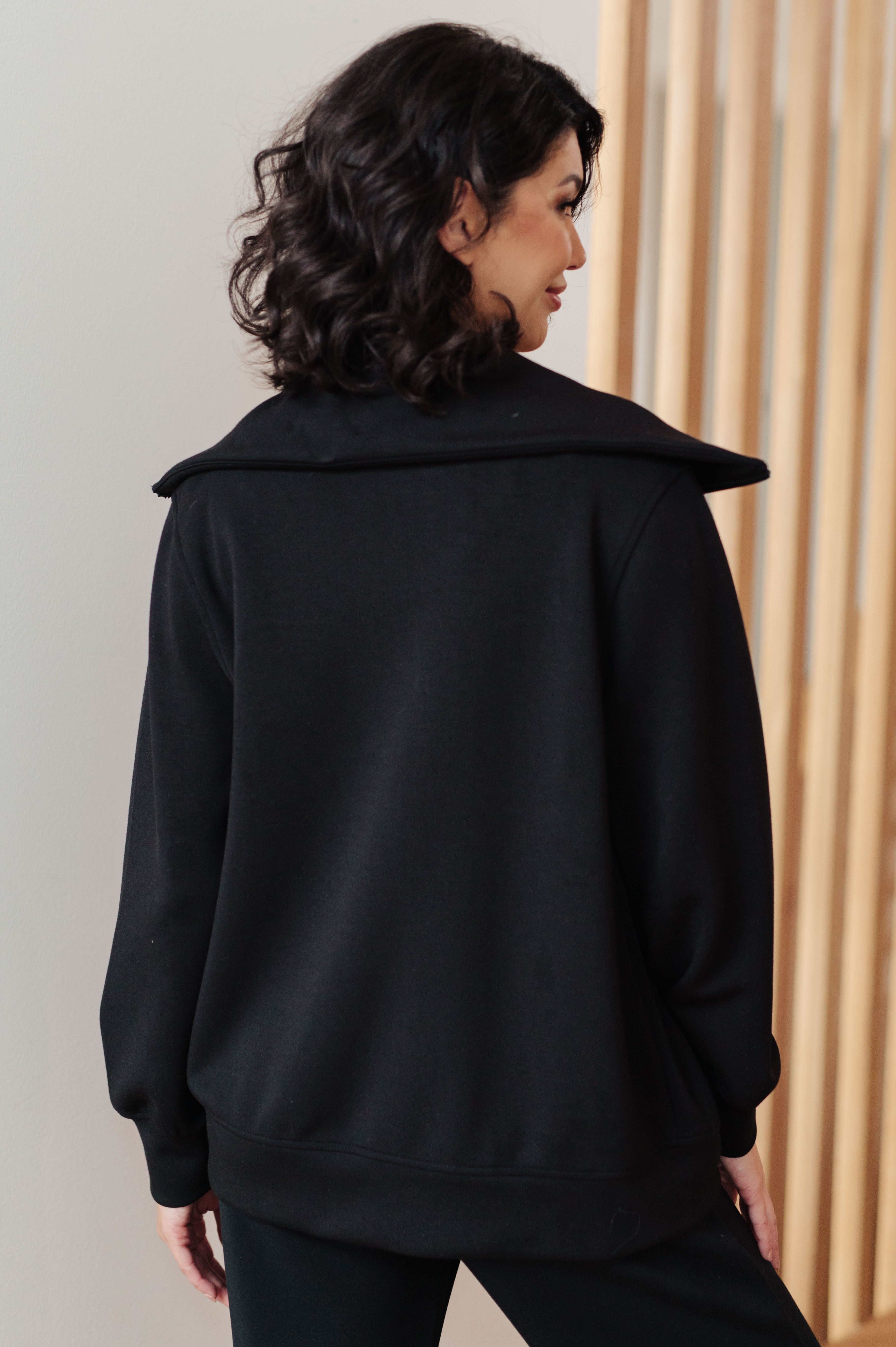 Woman wearing a athleisure black high neck zip up jacket and black pants standing in an indoor setting.