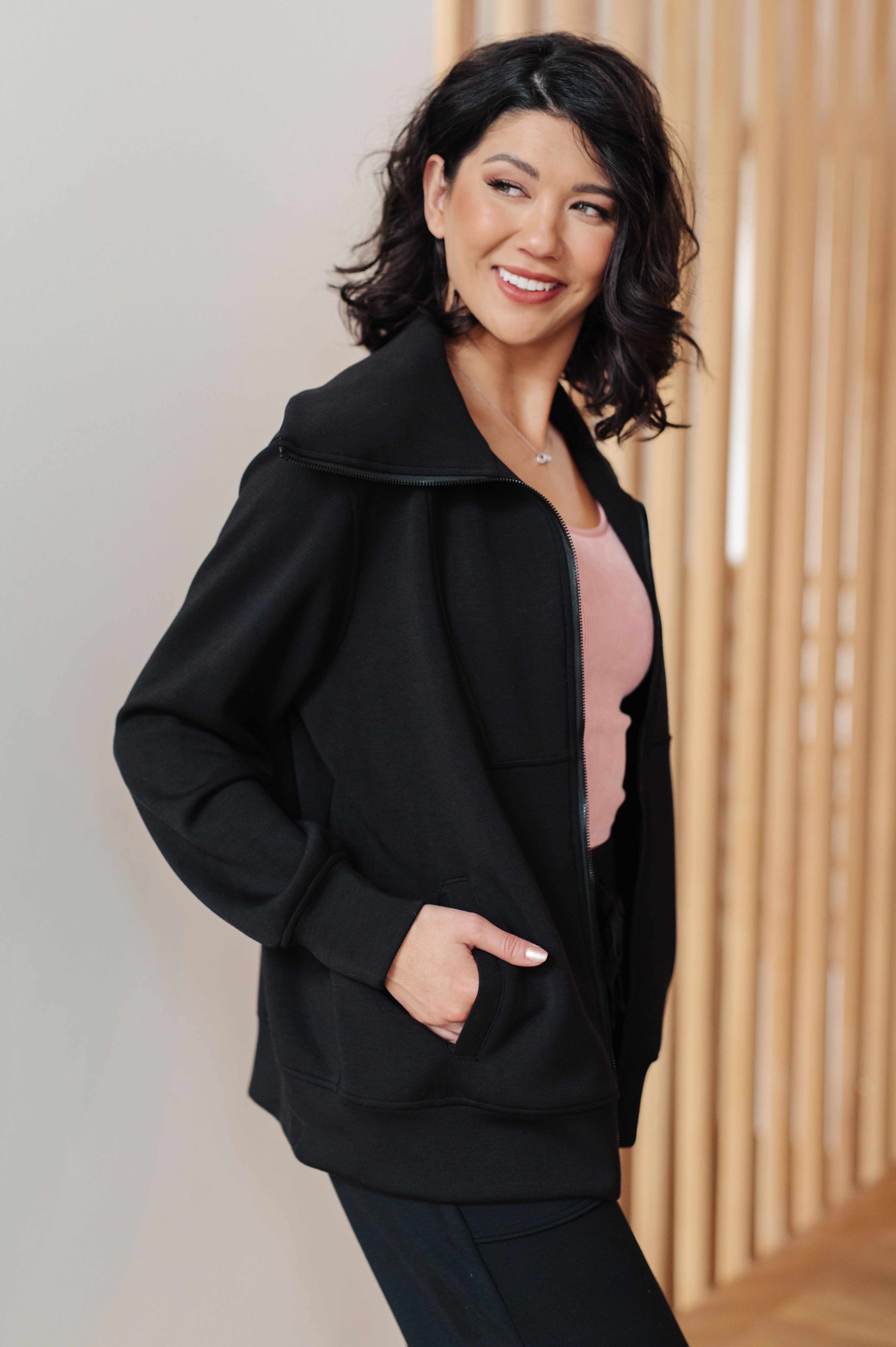 Woman wearing a athleisure black high neck zip up jacket, a pink top and black pants standing in an indoor setting.