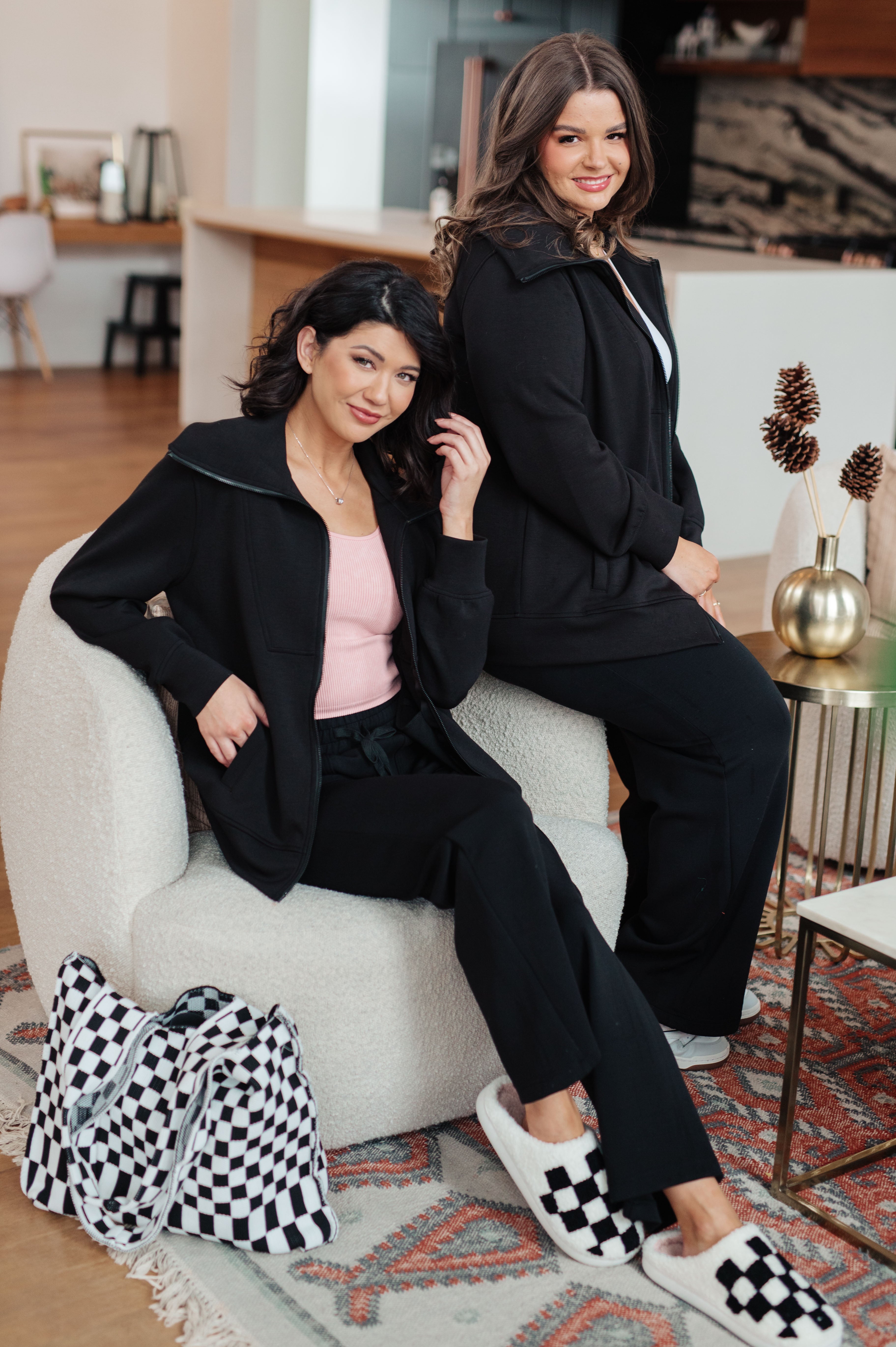 Two women in black athleisure outfits sitting on a white chair in a living room.