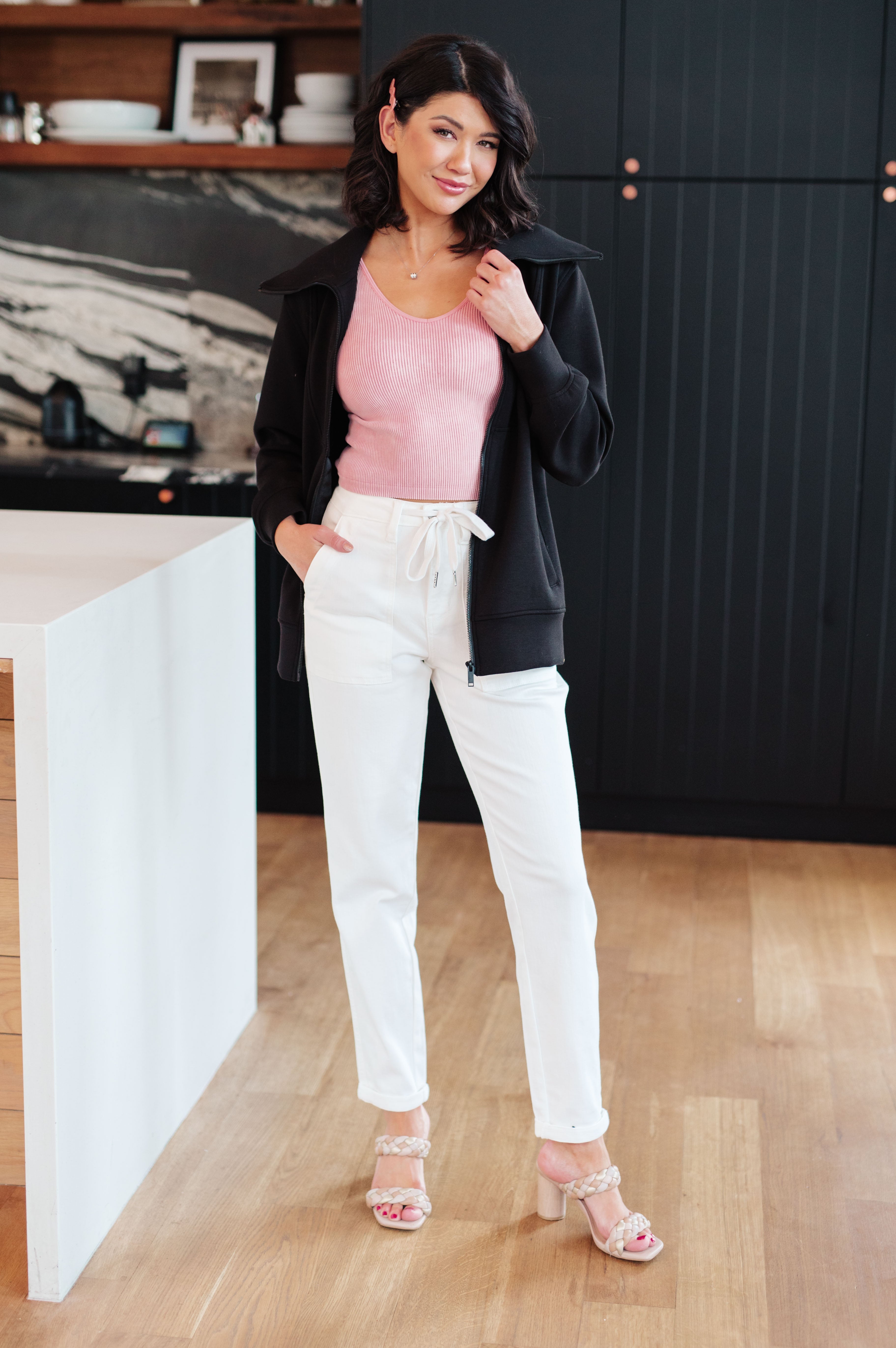 Woman wearing a athleisure black high neck zip up jacket, a pink top, white pants and beige sandals standing in an indoor setting.