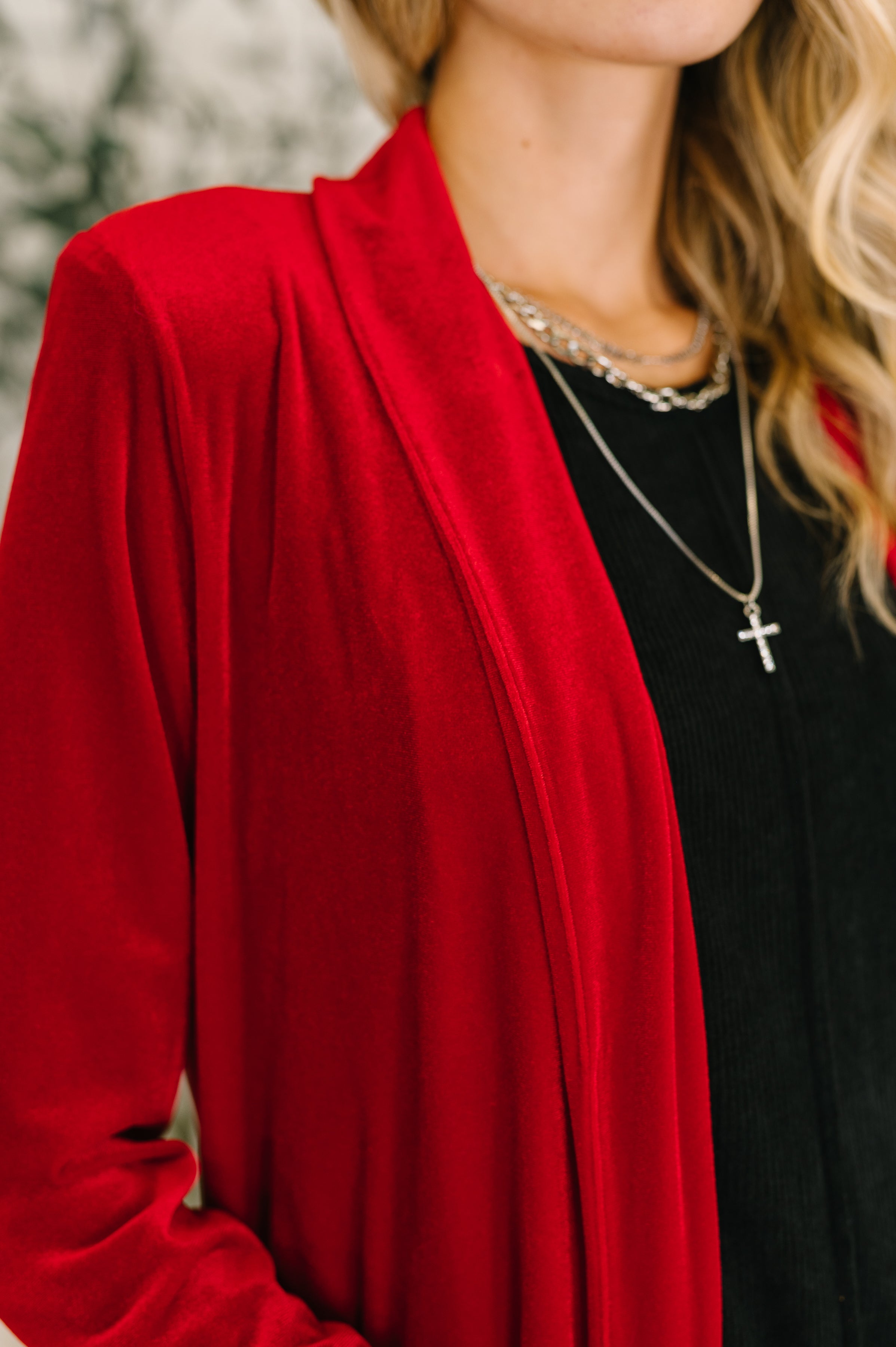 A close up of woman wearing a red velvet cardigan, a black dress and gold necklaces with a blurred background