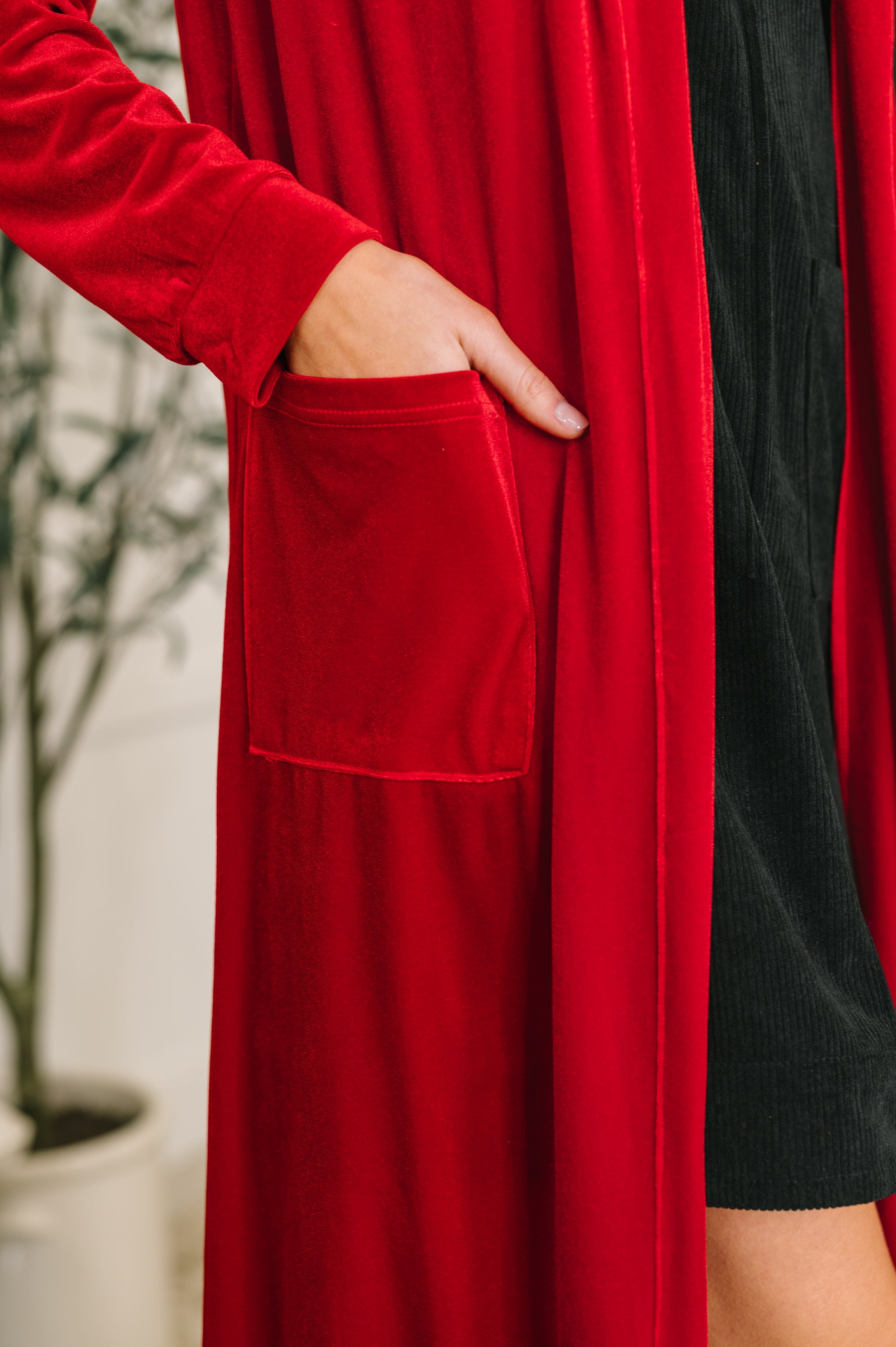 A close up of a woman wearing a red velvet cardigan over a black corduroy dress with a pocket against a neutral background.