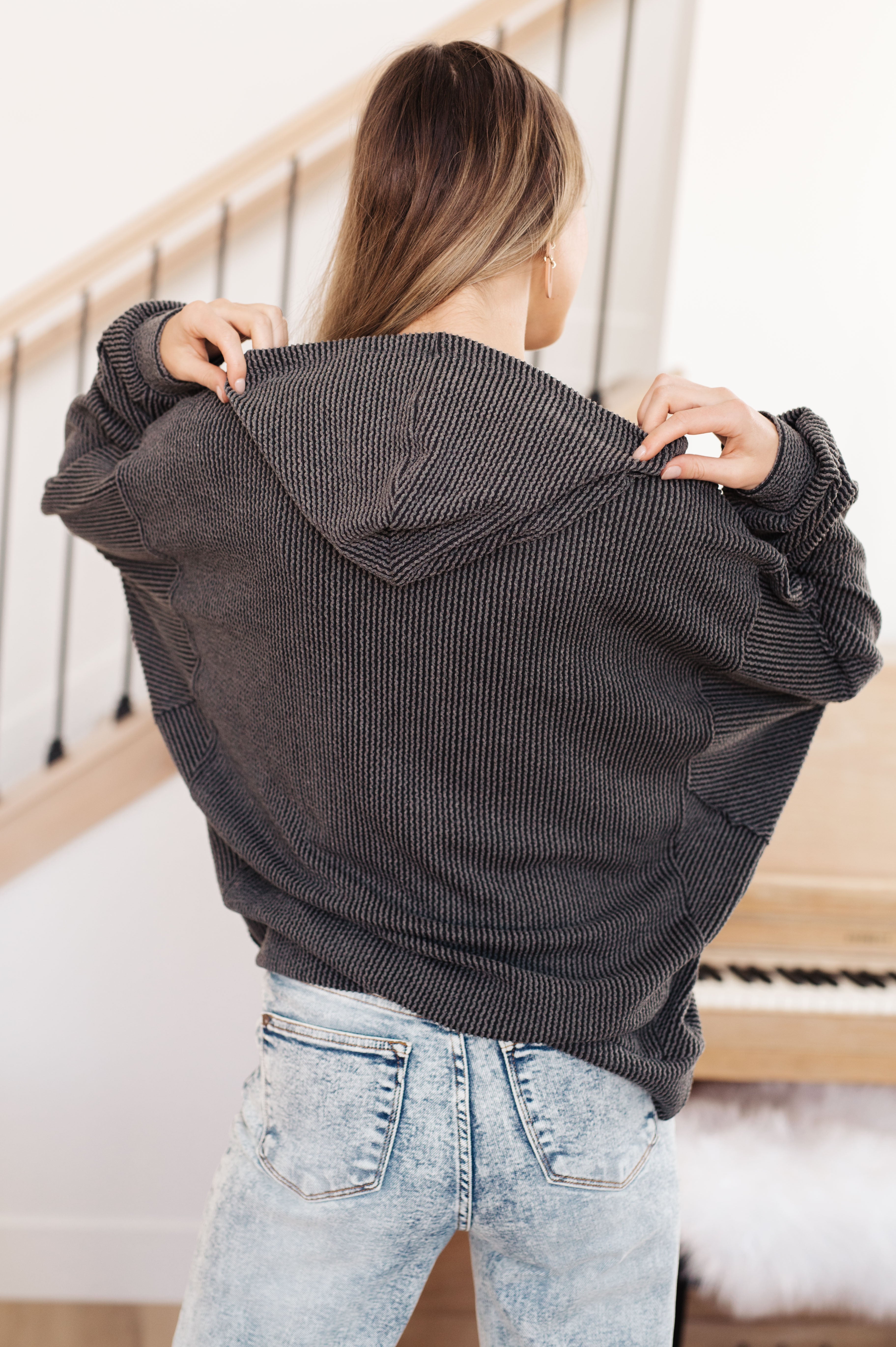 Woman with her back facing the camera wearing a grey rib knit hoodie over light blue jeans against an indoor setting.