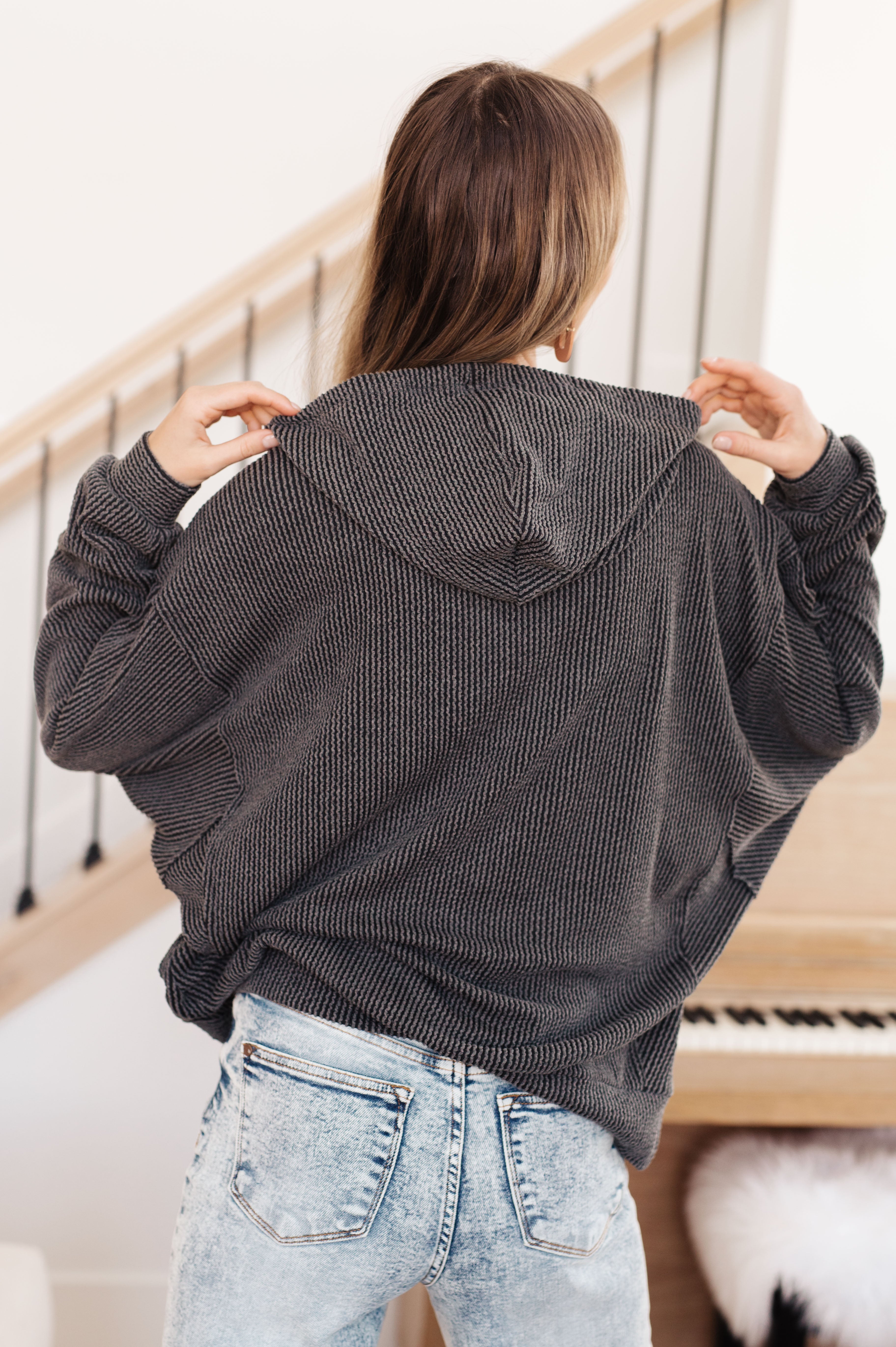 Woman with her back facing the camera wearing a grey rib knit hoodie over light blue jeans against an indoor setting.