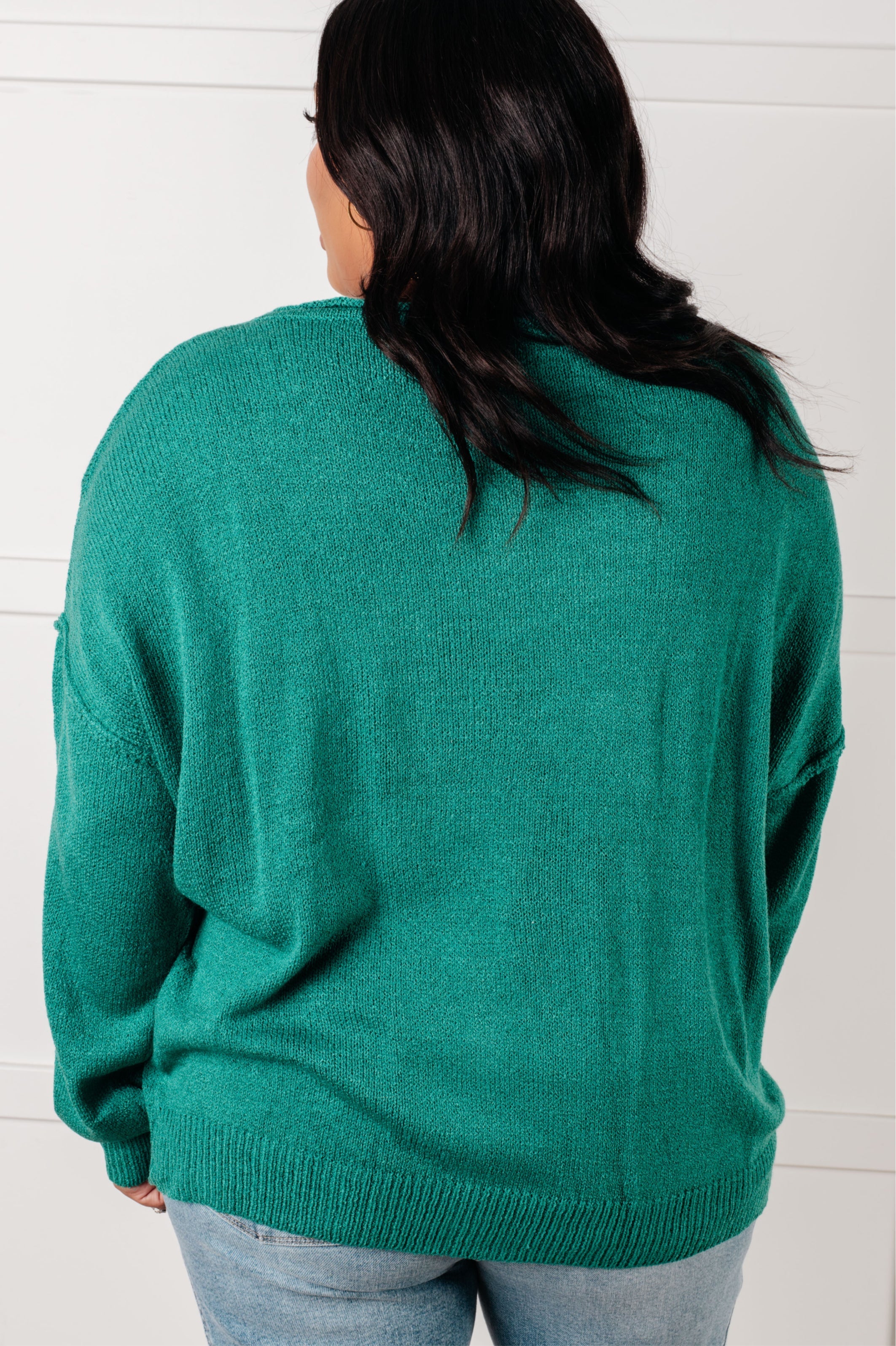 Woman with her back facing the camera wearing a teal sweater against over blue jeans against a white background.