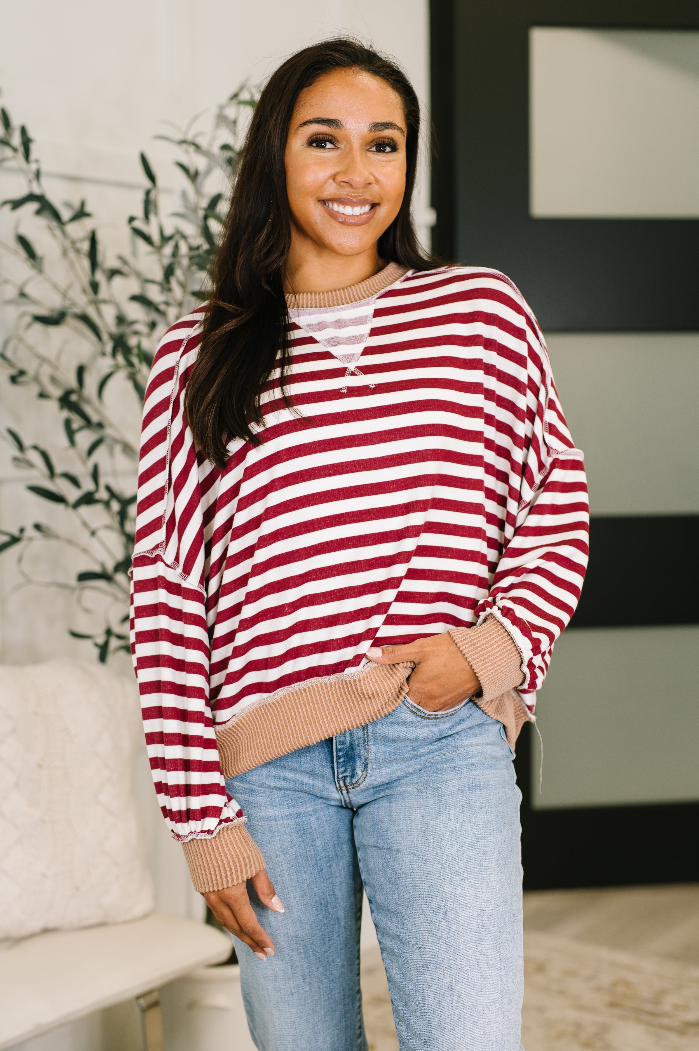 Woman wearing a red and white striped dolman sleeve pullover top with beige trim and blue jeans standing in an indoor setting.
