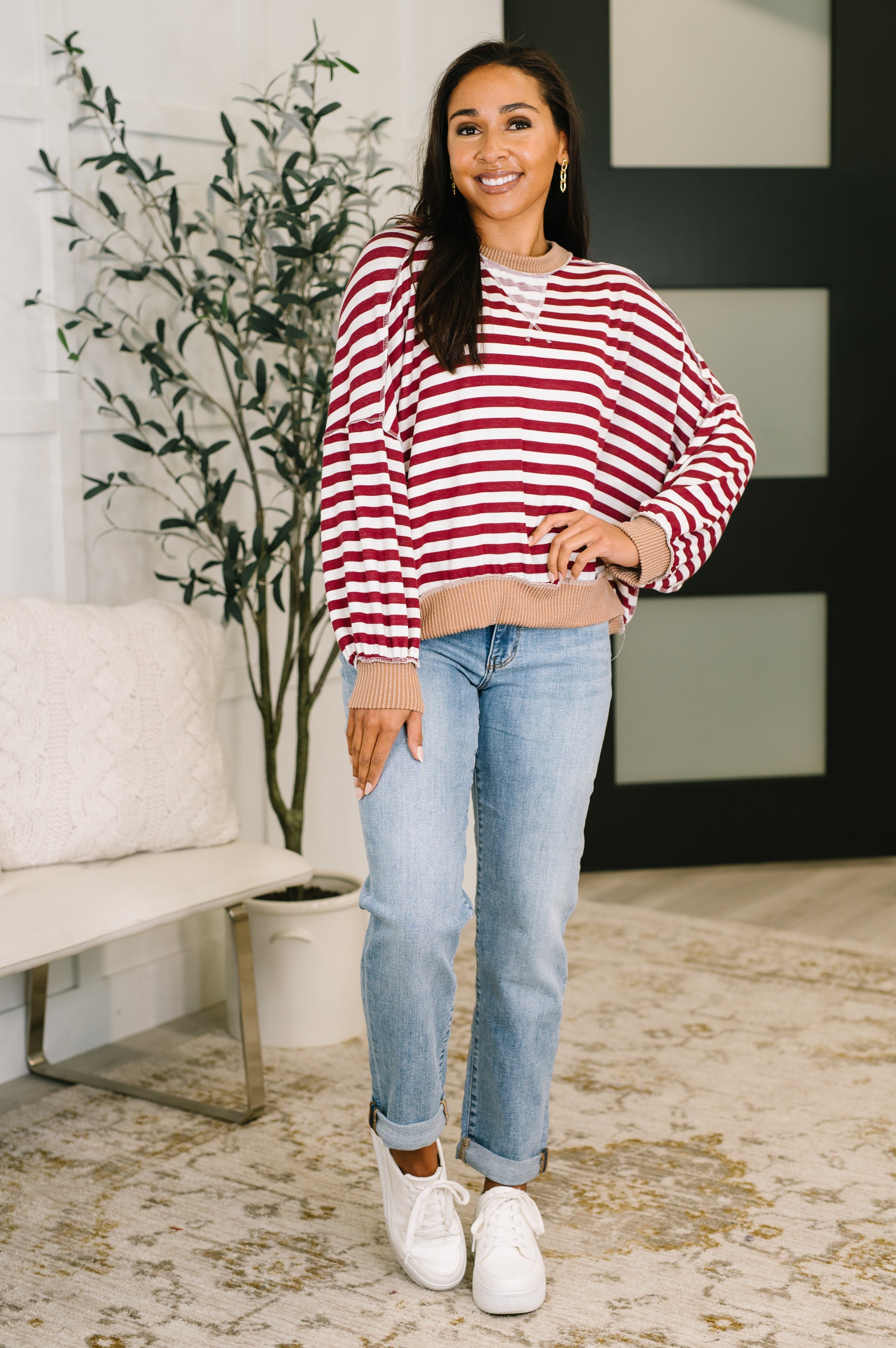 Woman wearing a red and white striped dolman sleeve pullover top with beige trim, blue jeans and white sneakers standing in an indoor setting.