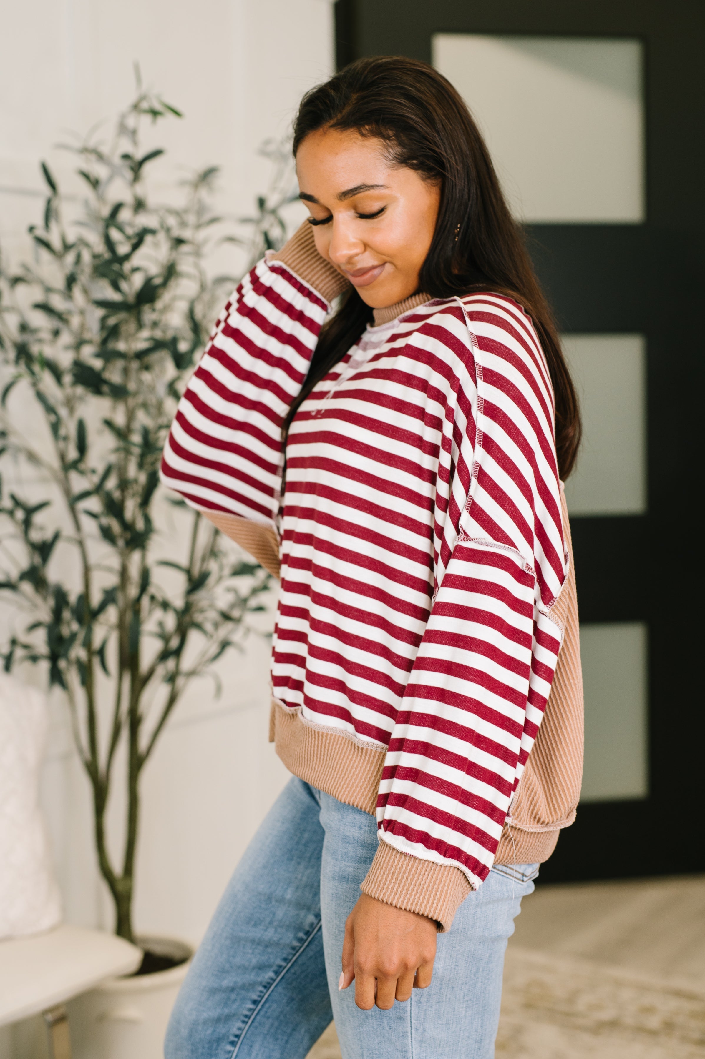 Woman wearing a red and white striped dolman sleeve pullover top with beige trim and blue jeans standing in an indoor setting.