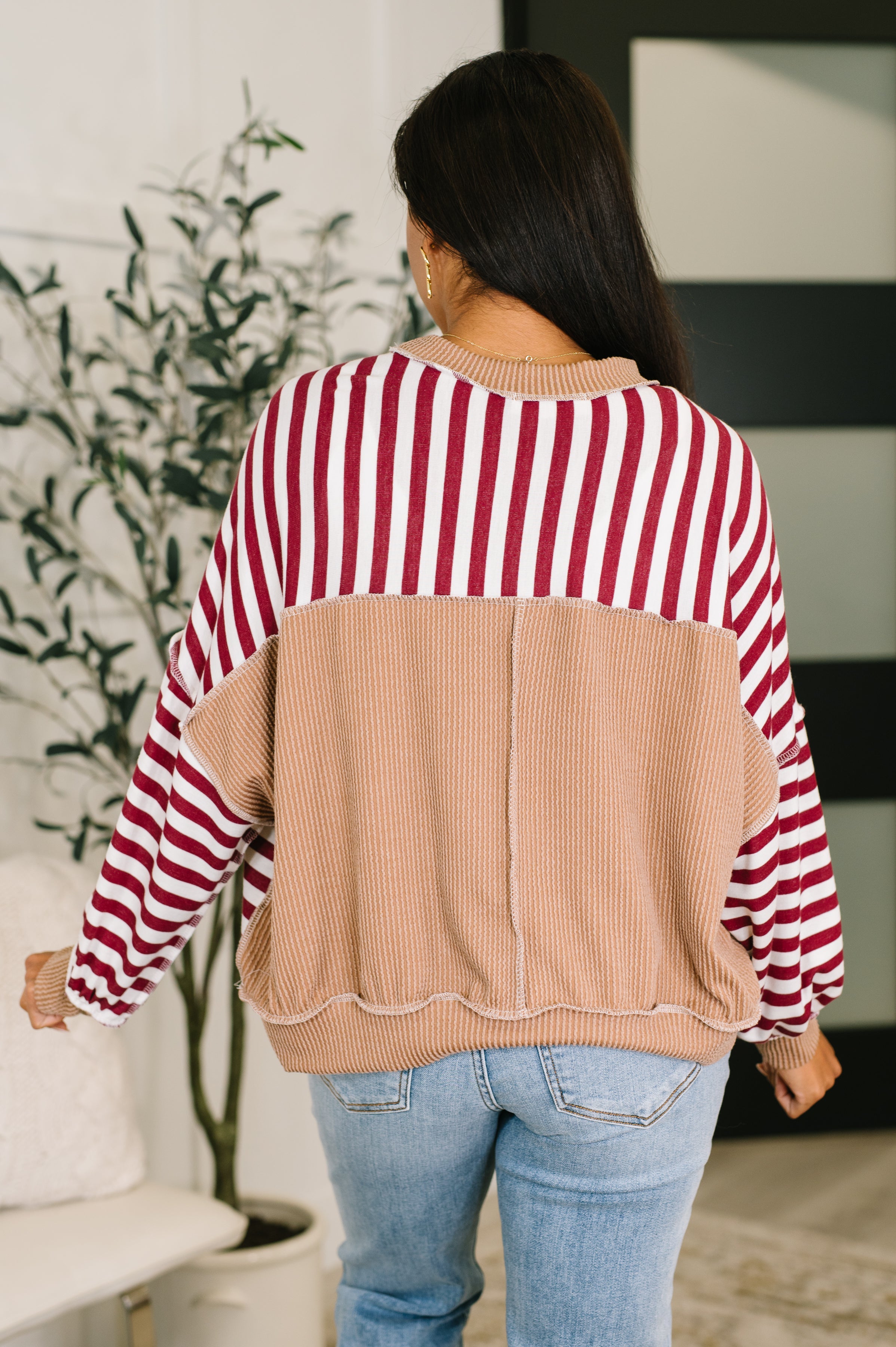 Woman wearing a red and white striped dolman sleeve pullover top with beige trim and blue jeans standing in an indoor setting.