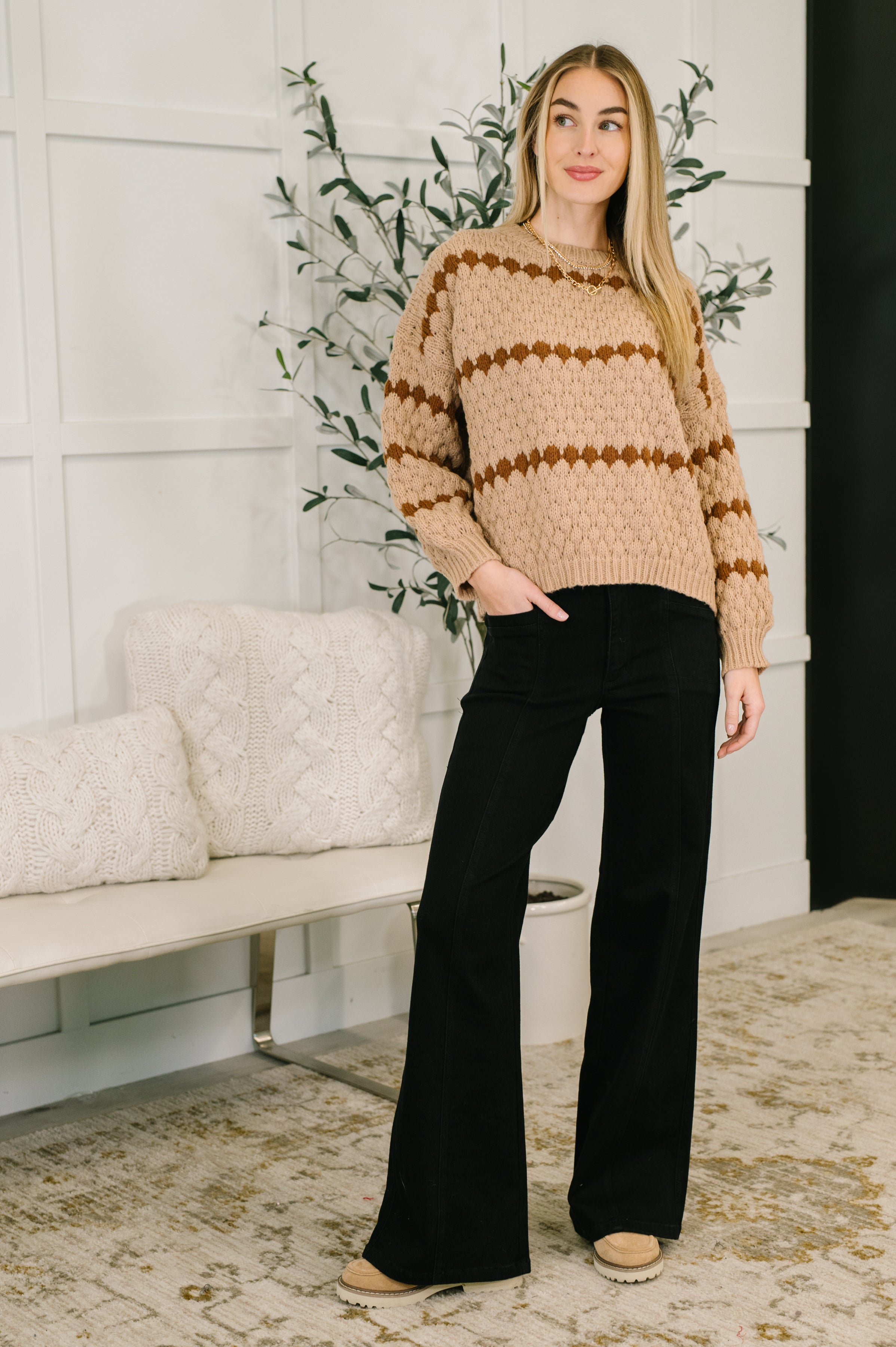 Woman wearing a chunky taupe sweater with brown stripes, black pants and brown boots in a living room setting.