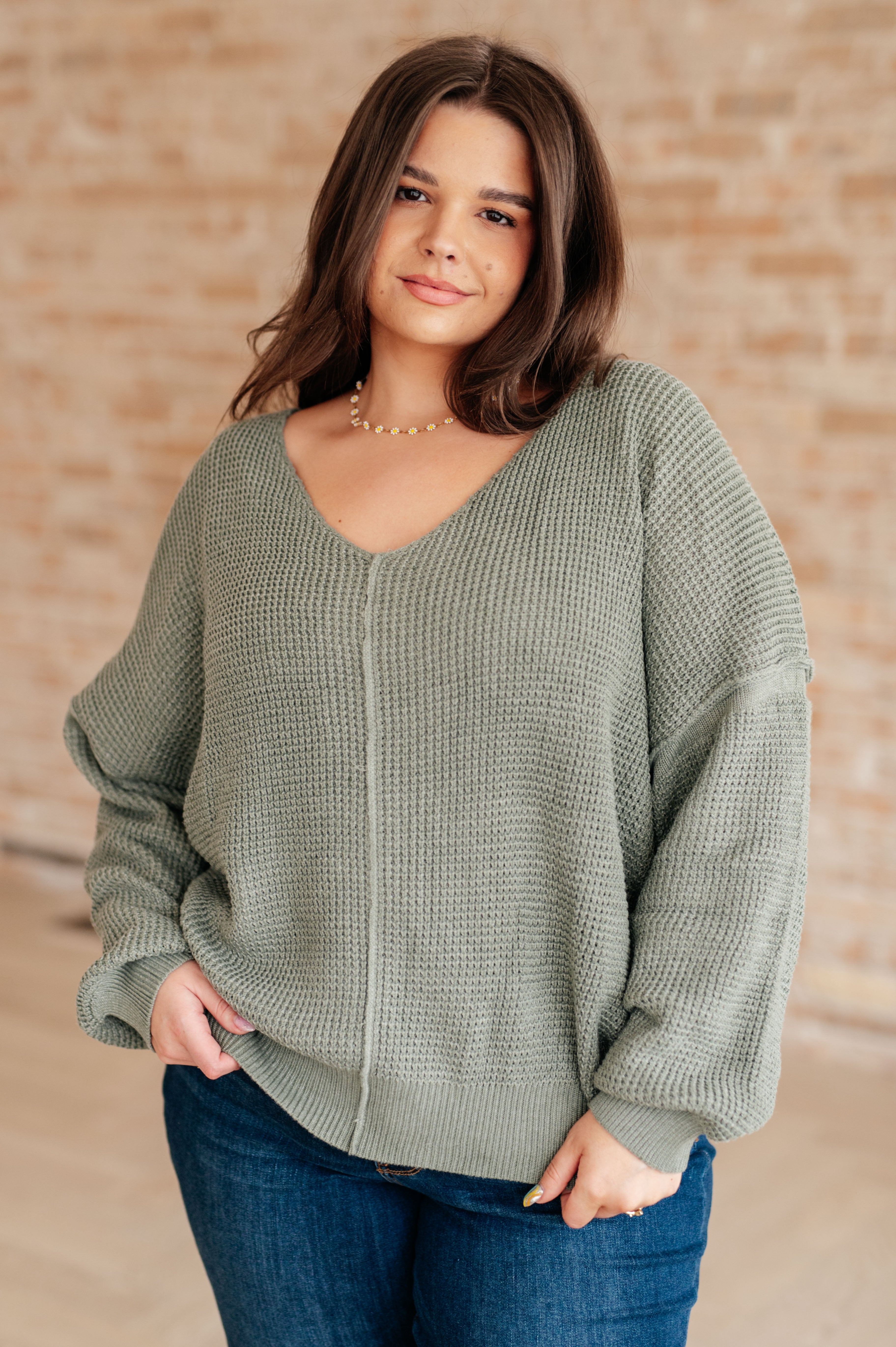 Woman wearing an olive green v-neck sweater with exposed seams and blue jeans standing against a brick wall