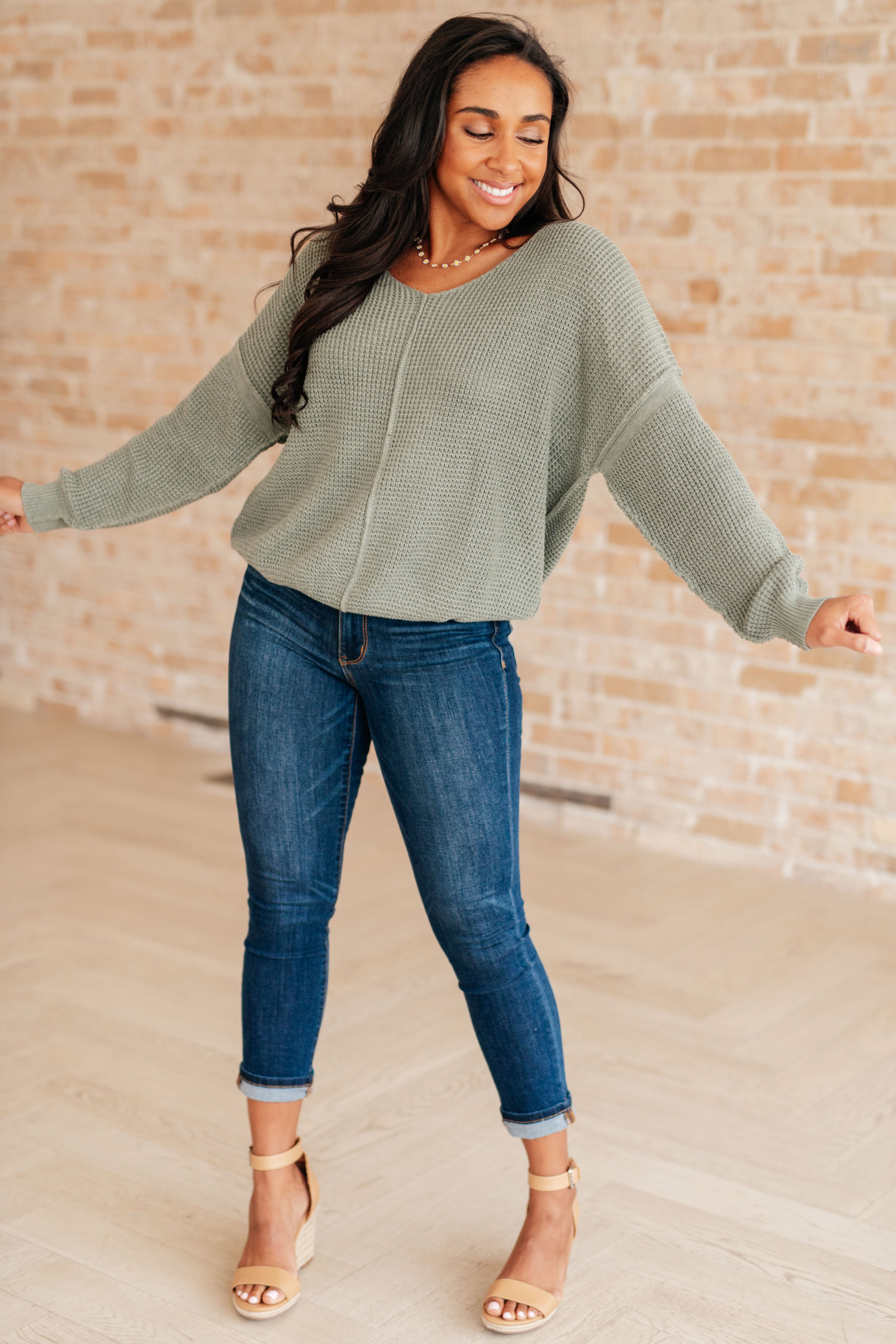 Woman wearing an olive green v-neck sweater with exposed seams, blue jeans and beige sandals standing against a brick wall