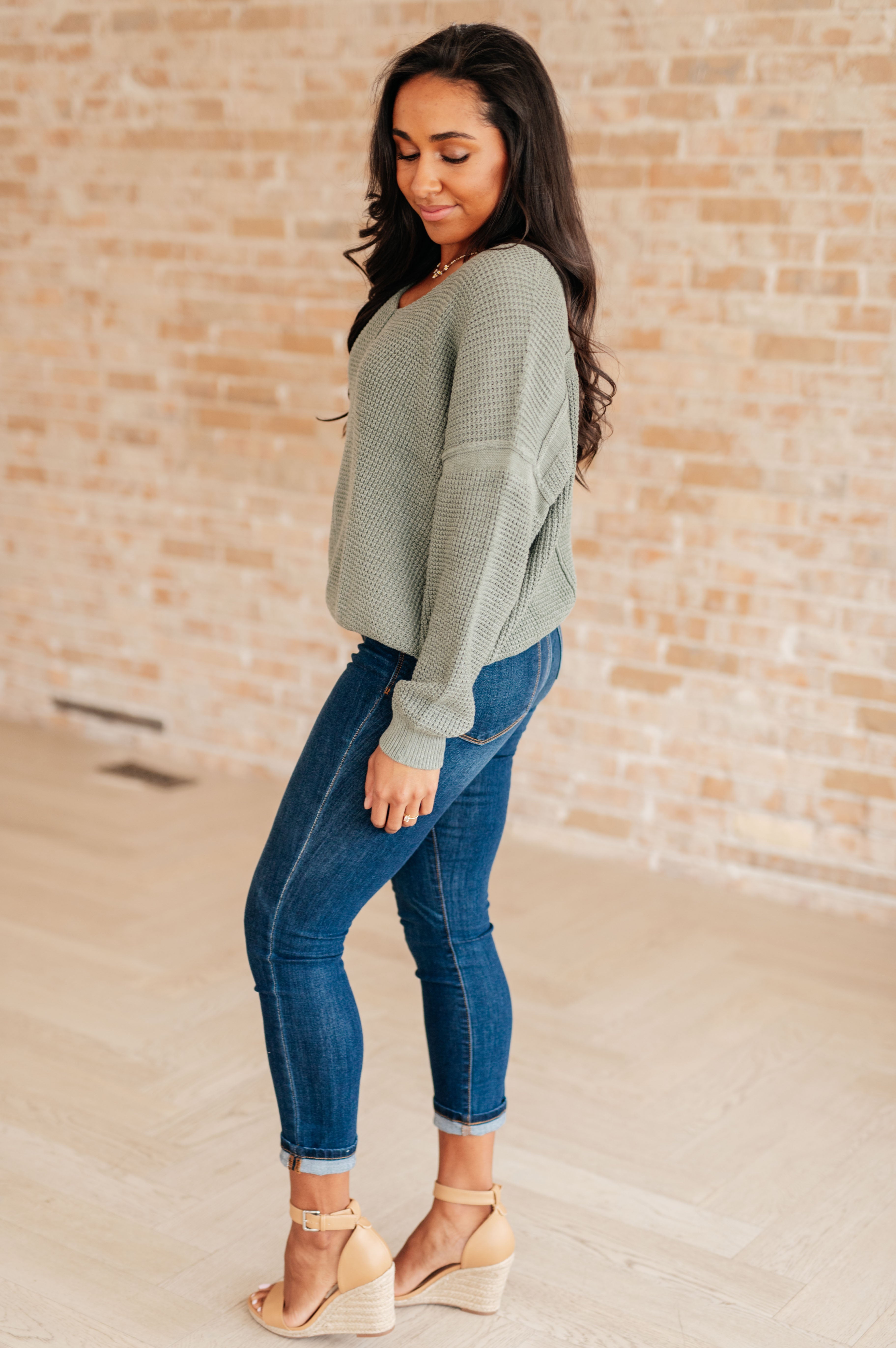 Woman wearing an olive green v-neck sweater with exposed seams, blue jeans and beige sandals standing against a brick wall