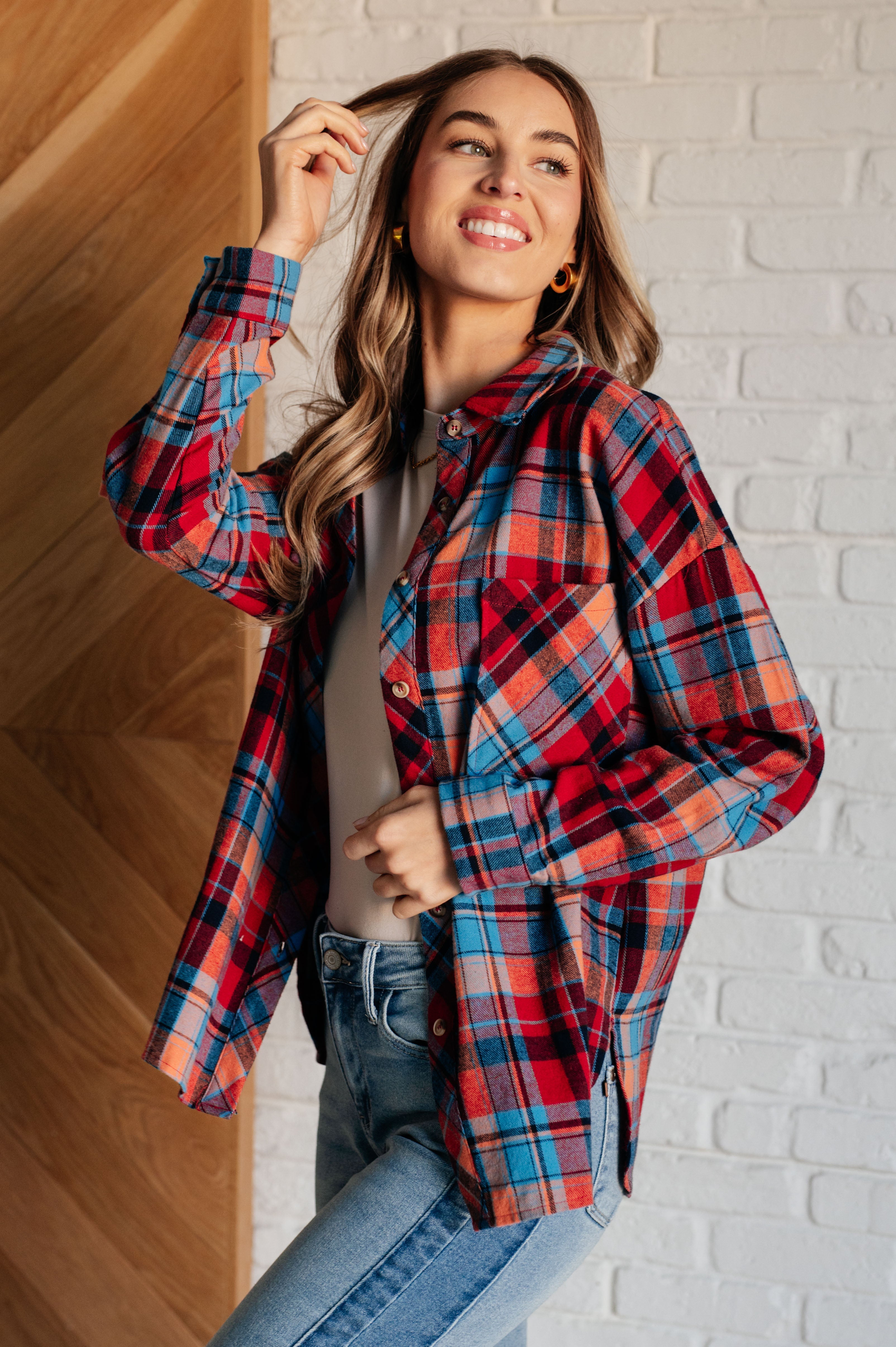 Woman wearing a red and blue long-sleeves plaid button-up shirt, a beige top and blue jeans with a white brick wall background.
