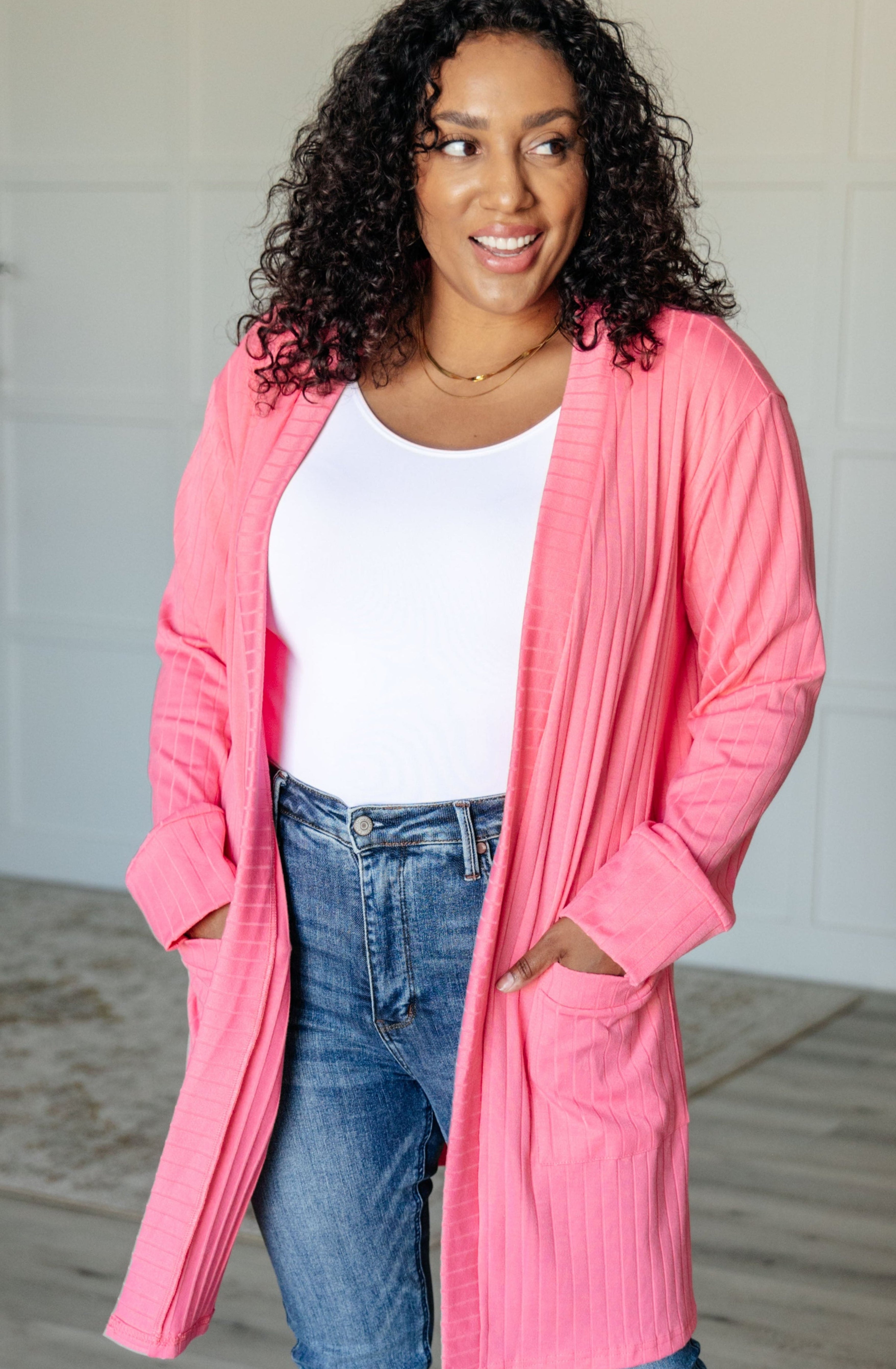 Smiling woman wearing bright pink ribbed open front Modern Idealist cardigan with pockets over white top and jeans indoors