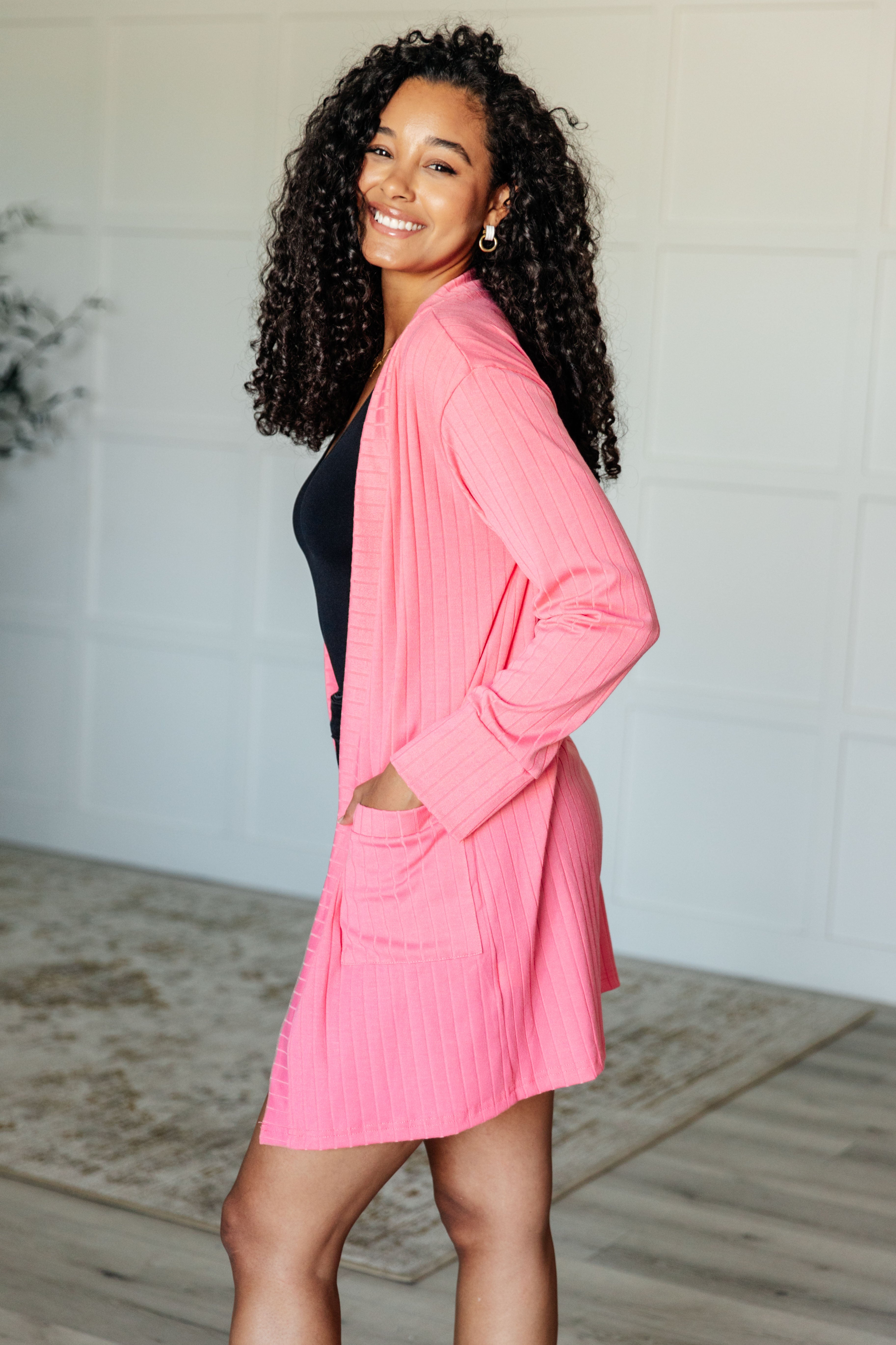 Smiling woman wearing bright pink ribbed Modern Idealist open front cardigan with pockets standing sideways indoors