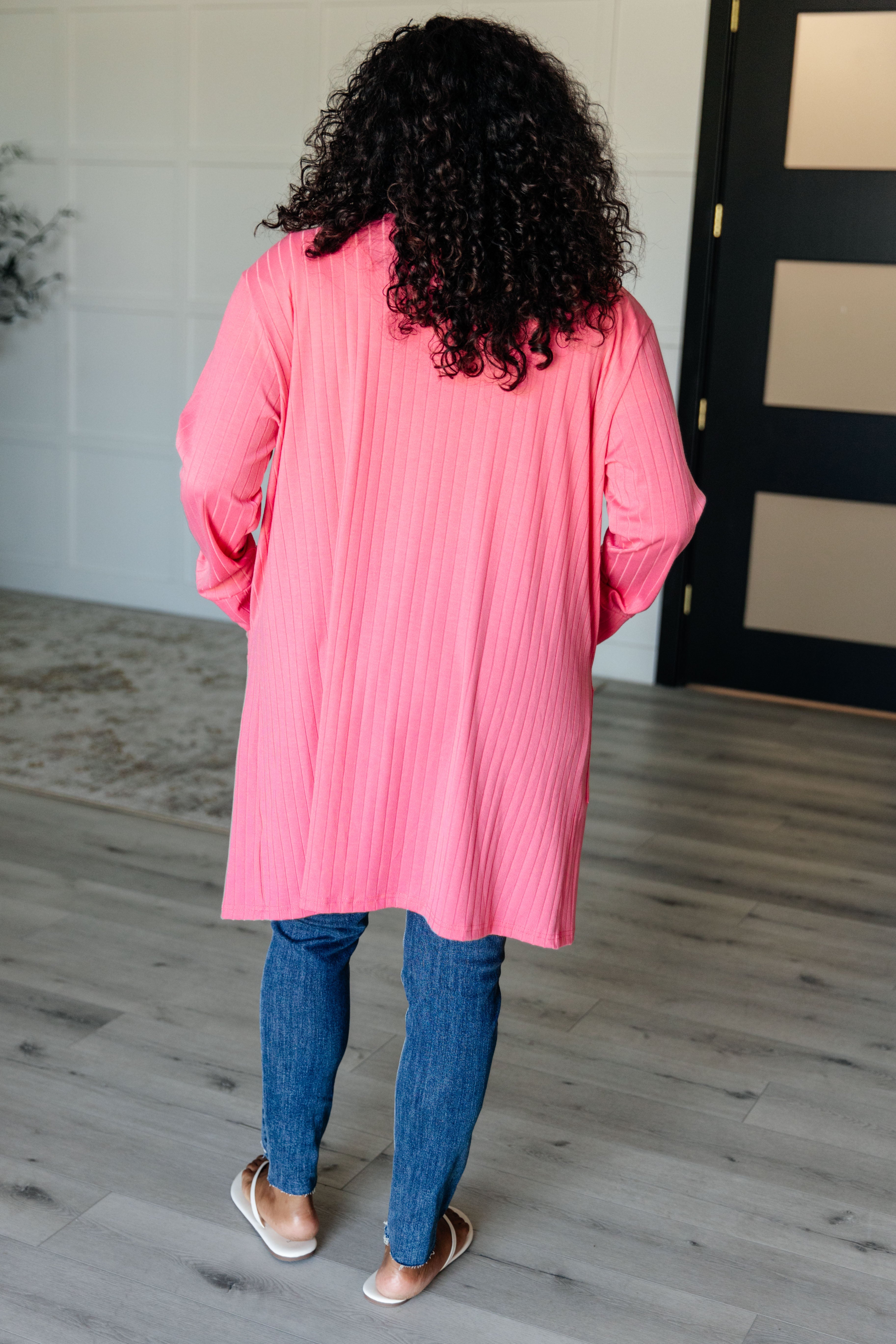 Woman seen from behind wearing bright pink ribbed open front Modern Idealist cardigan indoors on grey floor