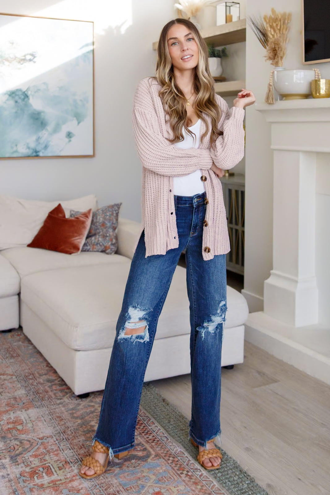 Woman wearing a light pink button down cardigan over a white top, distressed blue jeans and sandals standing in an indoor setting.