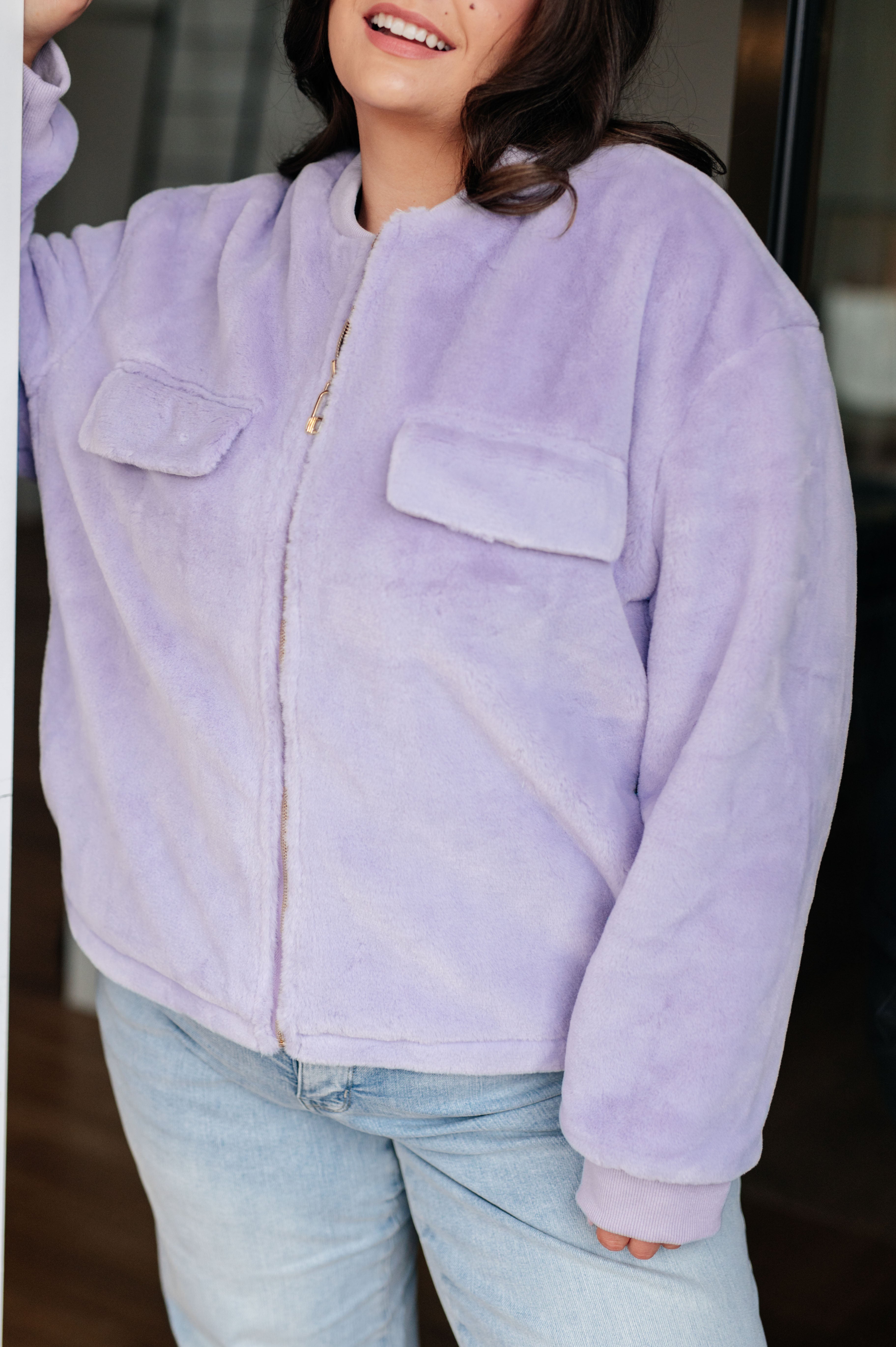 Woman wearing a light purple fuzzy zip up cropped coat and light blue jeans standing in front of a glass door.