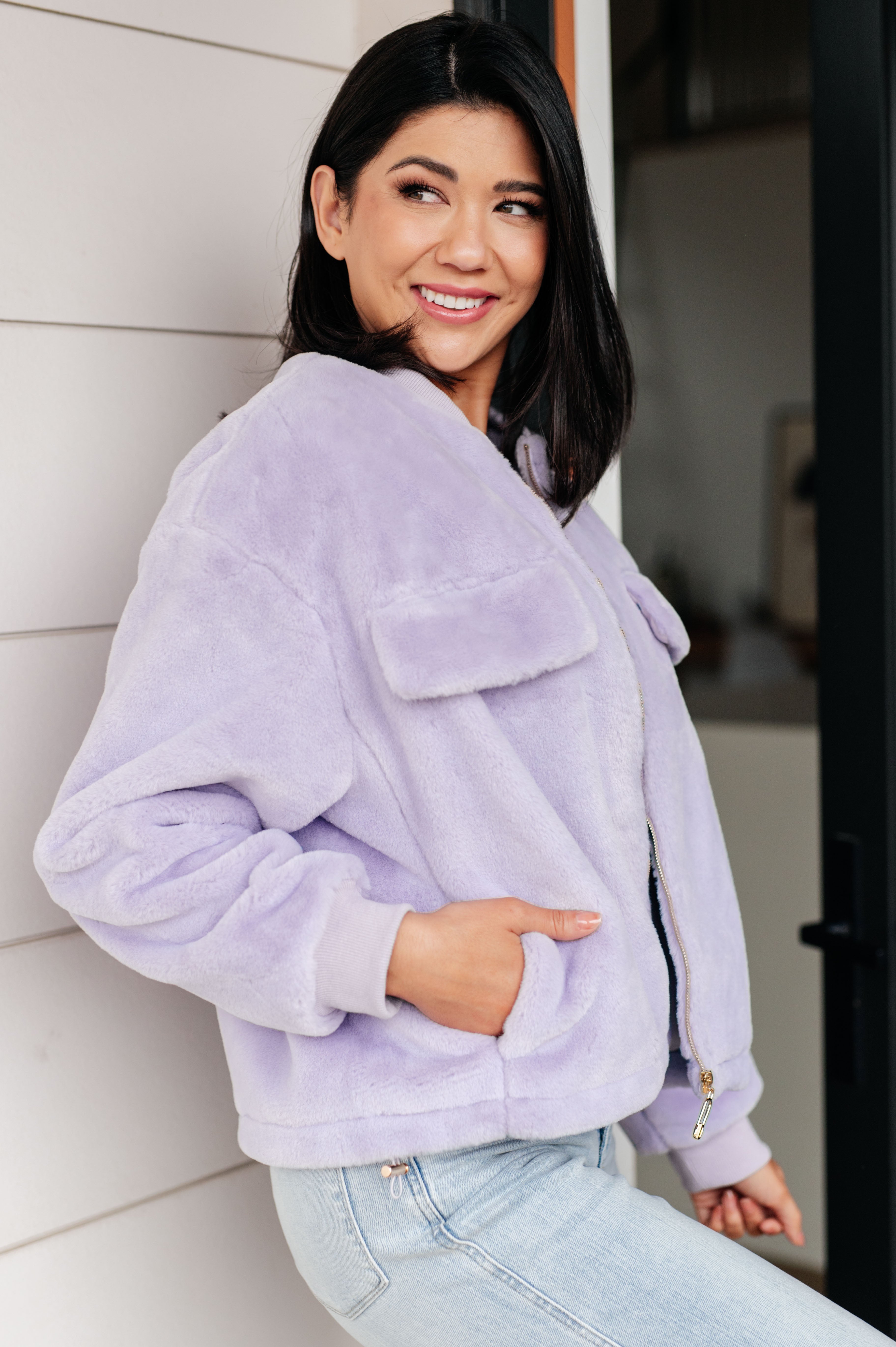 Woman wearing a light purple fuzzy zip up cropped coat and light blue jeans standing in front of a glass door.