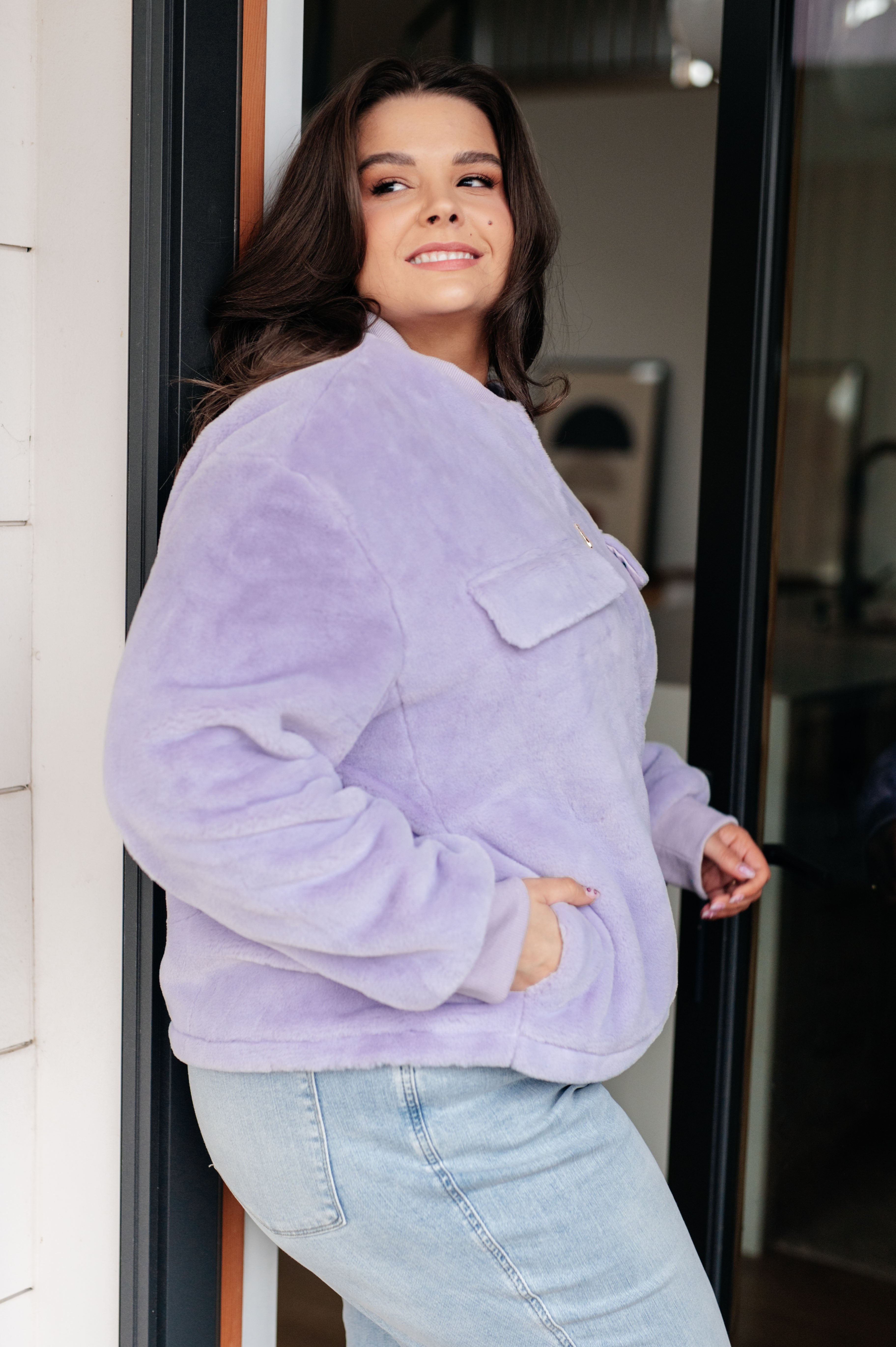 Woman wearing a light purple fuzzy zip up cropped coat and light blue jeans standing in front of a glass door.